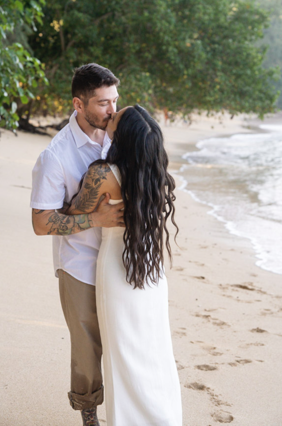 A couple kissing on a beach, with trees in the background and footprints in the sand.