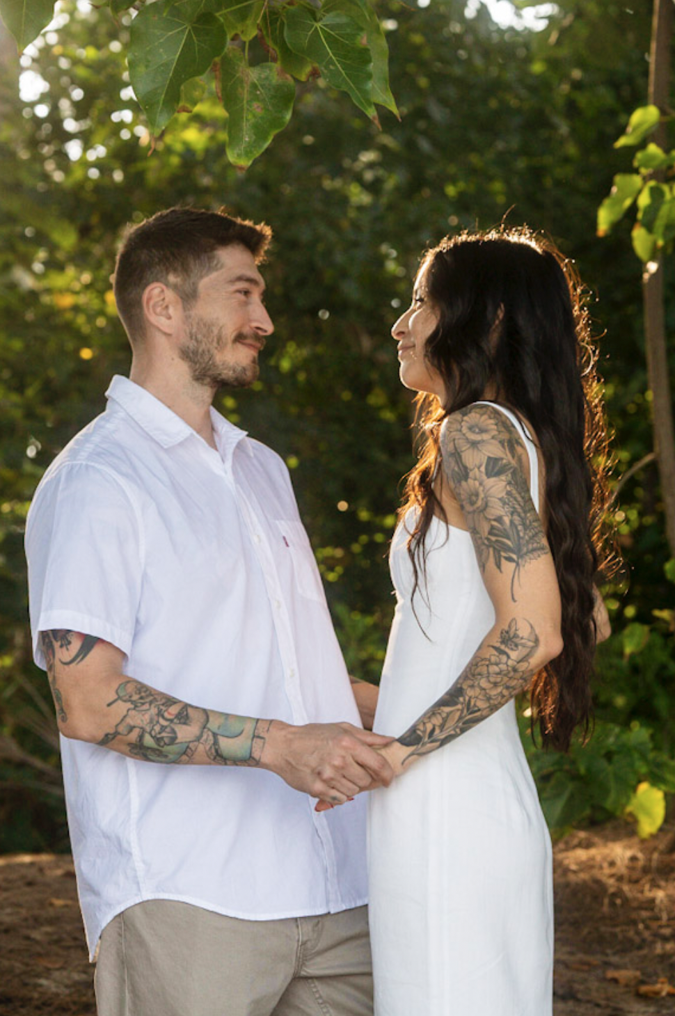 A couple holding hands and smiling at each other outdoors during sunset, surrounded by greenery.