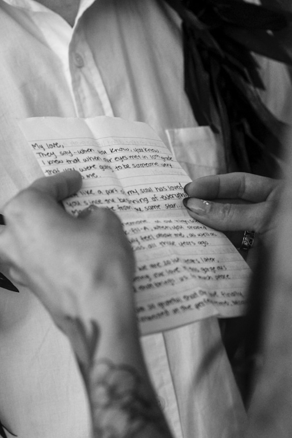 A person holding a handwritten letter, reading it. The person has a tattoo on their arm and is wearing a ring. The letter is written on lined paper and the person appears to be reading it carefully.