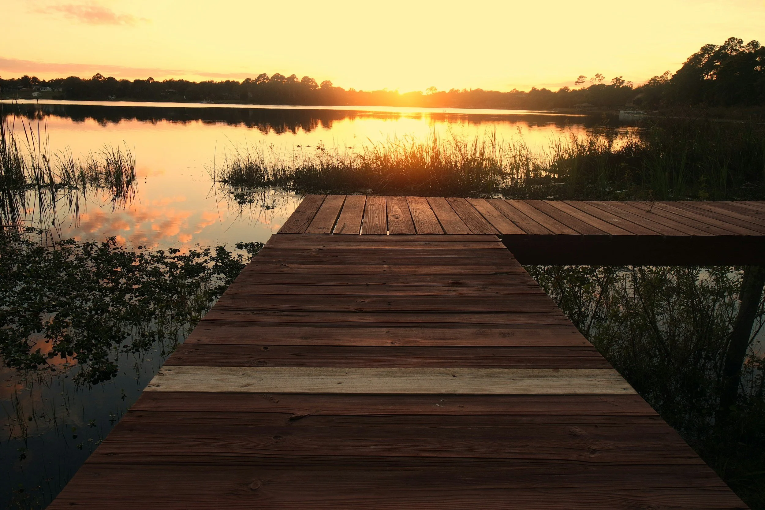 A wooden dock extends over a calm lake at sunset, with the sky glowing orange and yellow, and the landscape reflected in the water.
