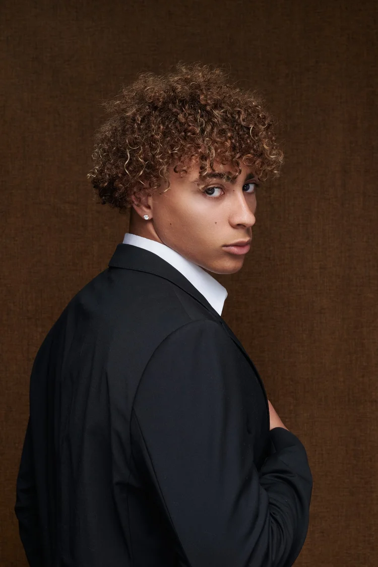 Portrait d'une jeune femme portant un costume noir et une chemise blanche, avec des cheveux bouclés bruns et une boucle d'oreille en diamant, regardant par-dessus son épaule sur un fond marron.