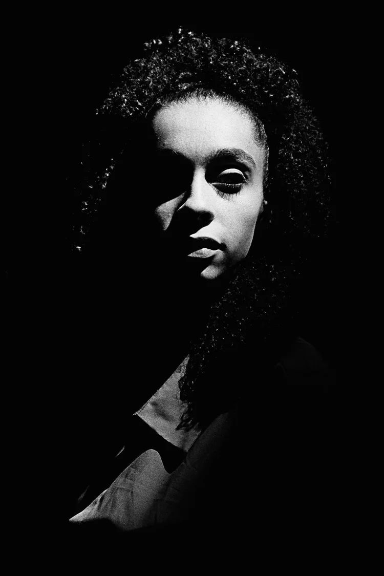 Portrait en noir et blanc d'une femme avec de longs cheveux bouclés, partiellement ombragée, portant un vêtement clair.