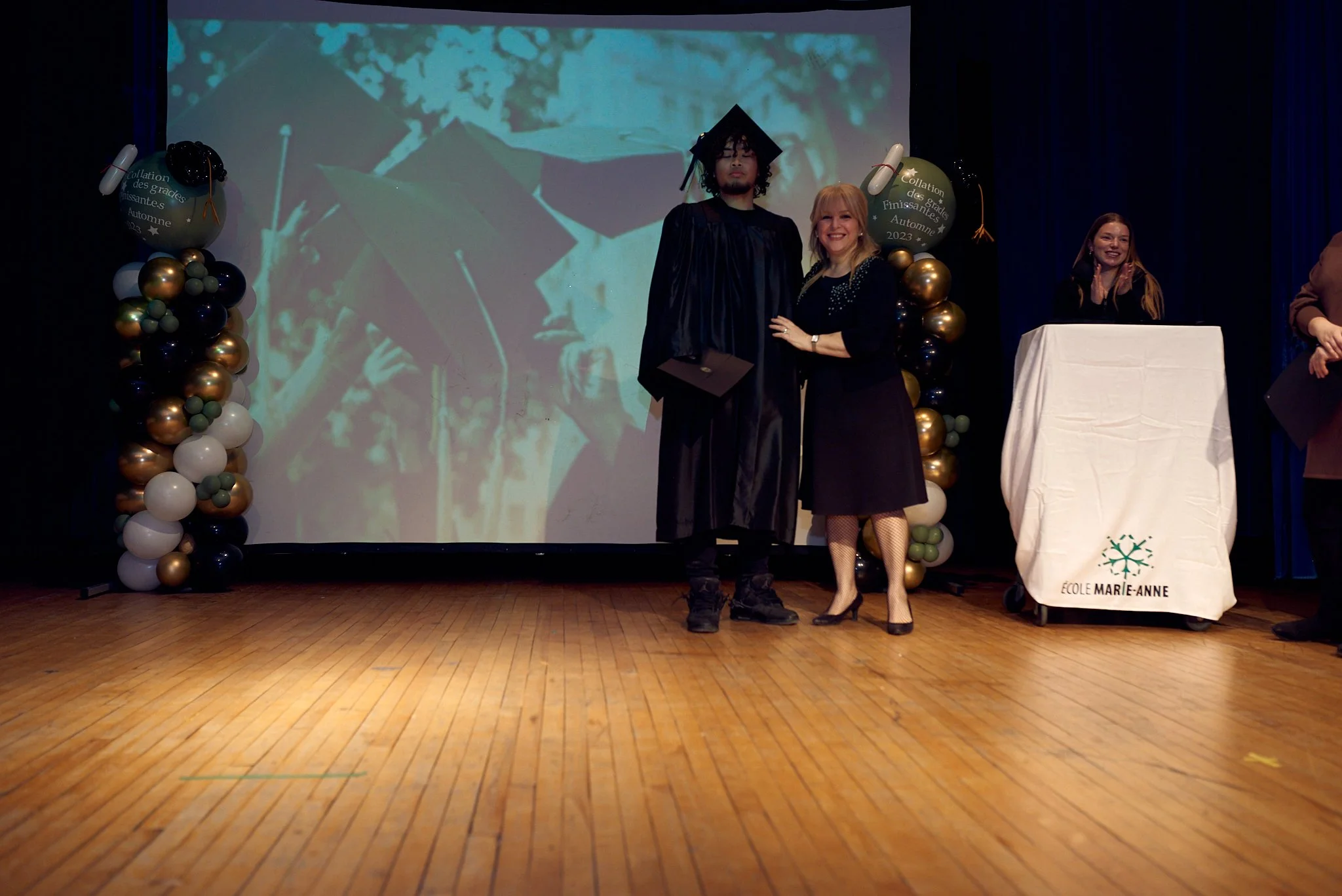 Un étudiant en graduation posant avec une femme lors d'une cérémonie de remise de diplôme à l'école Marie-Anne. Il porte une toge et un chapeau de graduation, tandis qu'elle porte une robe noire. La scène se déroule sur une scène avec un fond projeté