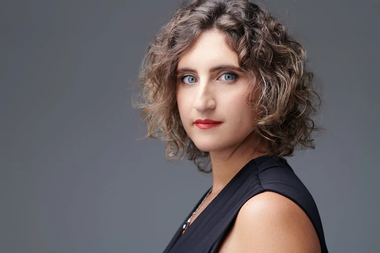 Portrait d'une femme avec des cheveux bouclés, yeux bleus, portant un vêtement noir, fond gris.