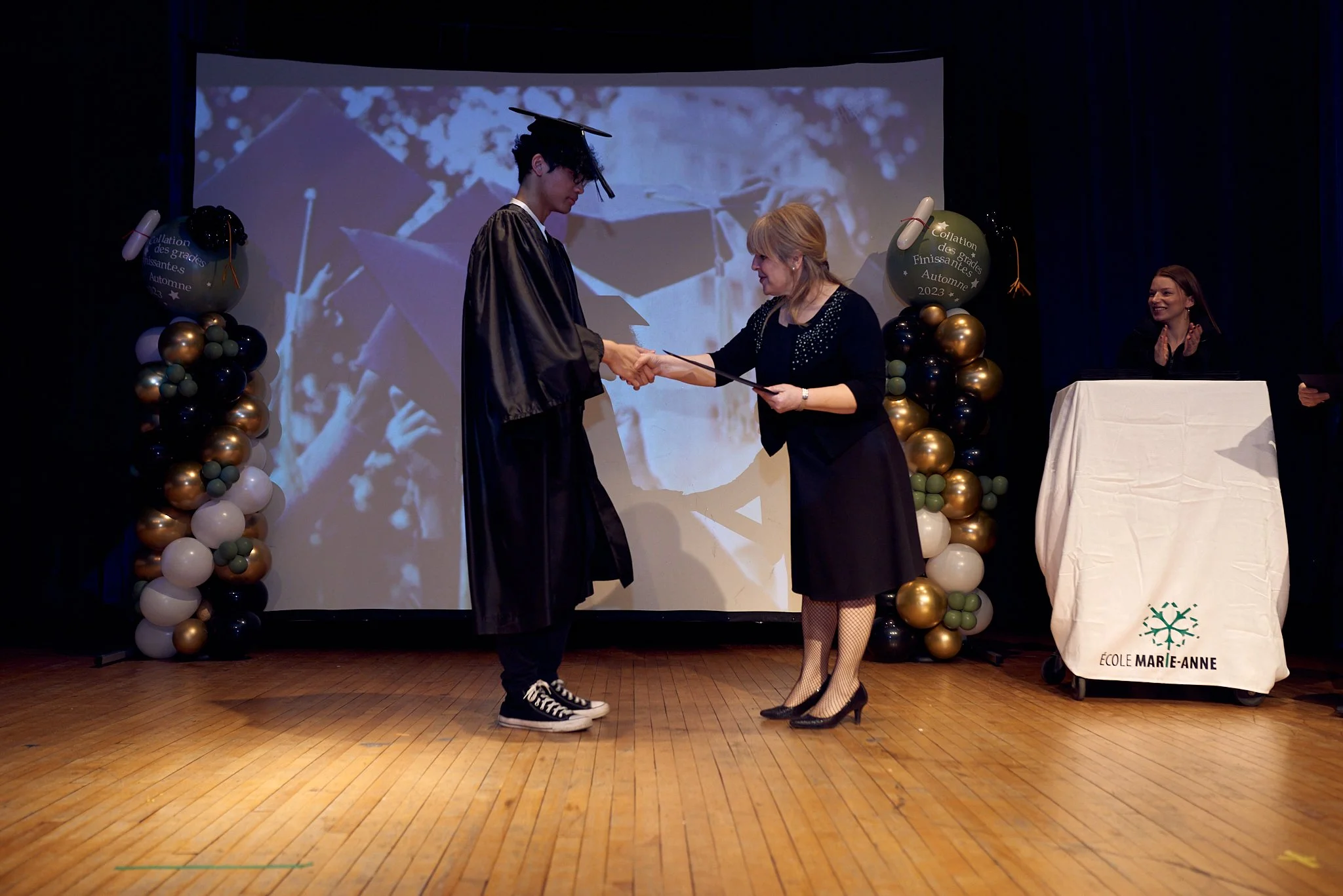 Un jeune homme en robes de diplômé, portant un chapeau de graduation, reçoit un diplôme d'une femme lors d'une cérémonie de remise de diplômes organisant à l'école Marie-Anne. Deux autres personnes sont visibles, l'une saluant à côté d'une table couv