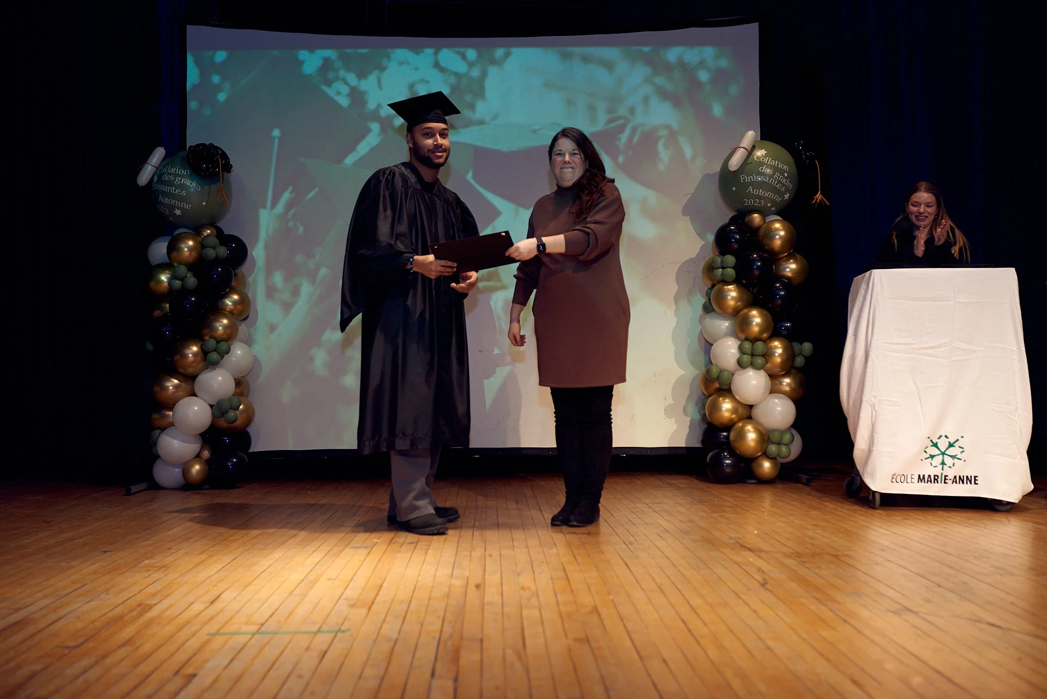 Un diplômé en robe de graduation recevant son diplôme d'une femme lors d'une cérémonie sur scène. Décor avec des ballons dorés, blancs et noirs, et une bannière indiquant l'école Marie-Anne.