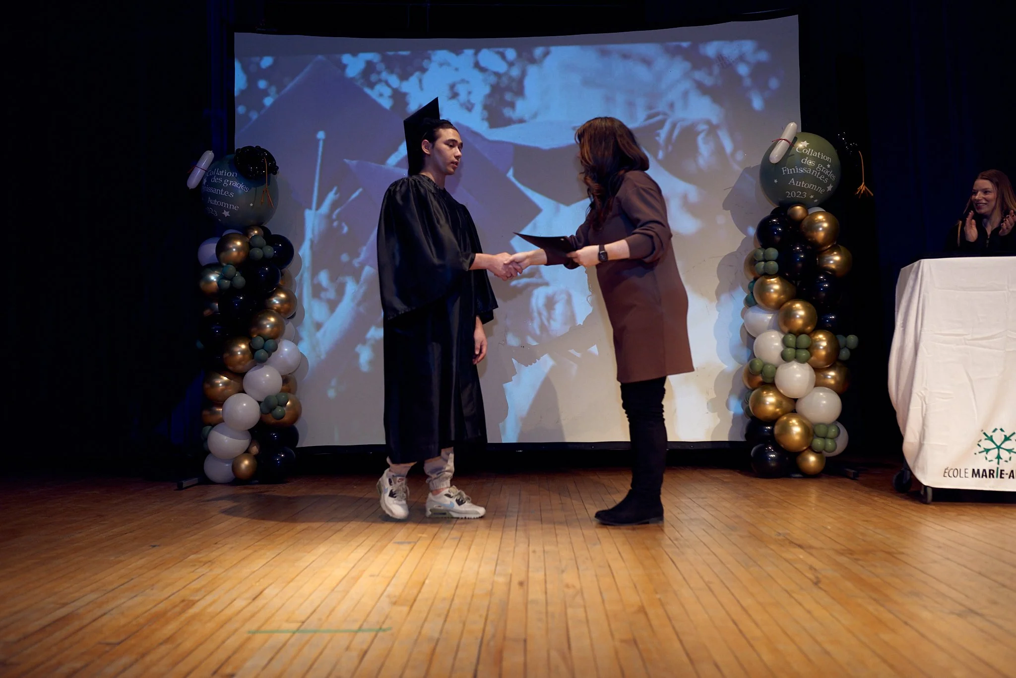 Un jeune homme en robe de graduation recevant un diplôme d'une femme lors d'une cérémonie, avec des ballons dorés, noirs, verts et blancs décorant le fond et une personne applaudissant à droite.