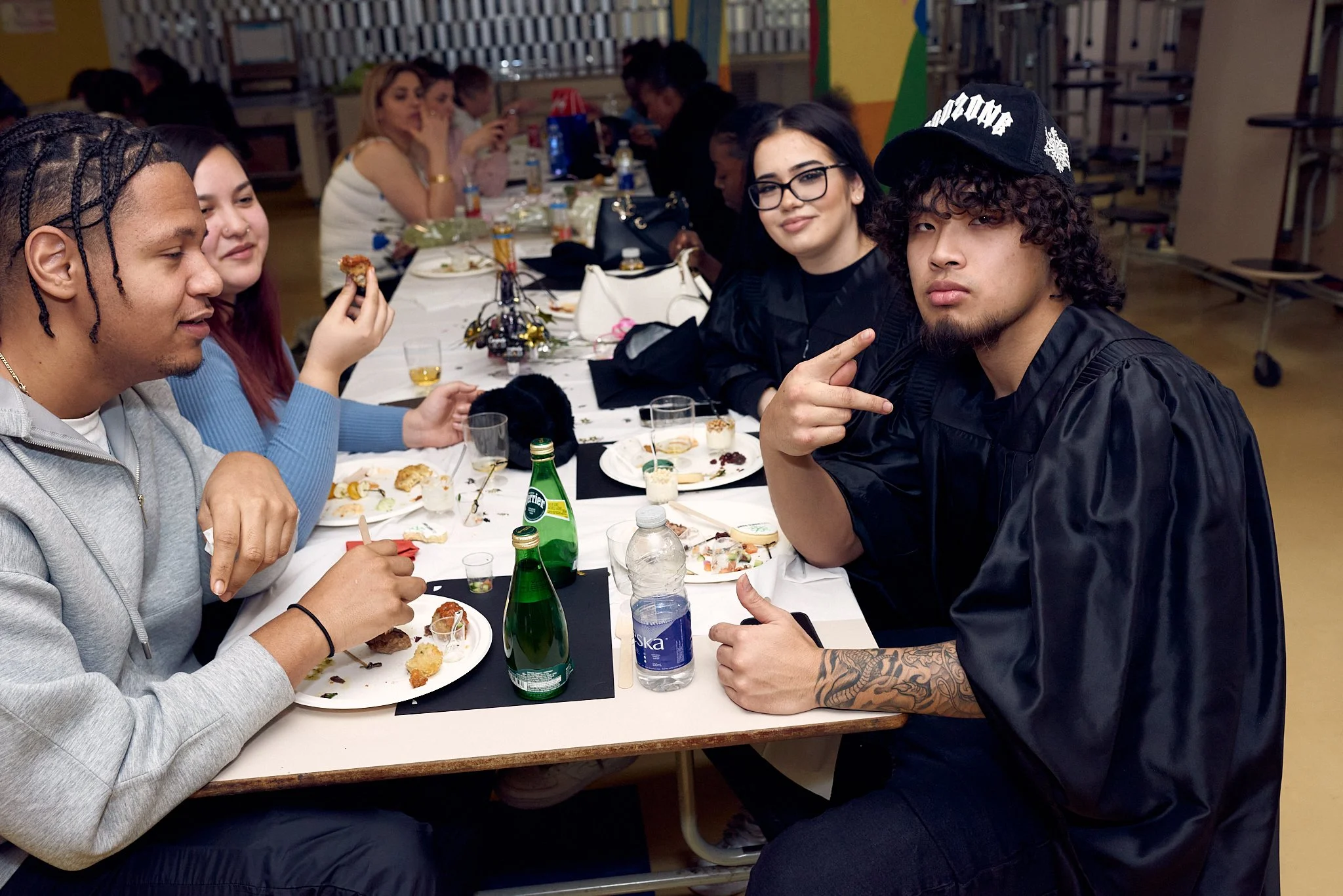 Groupe de jeunes adultes assis à une table mangeant et socialisant lors d'un repas, dans une salle communautaire ou scolaire.