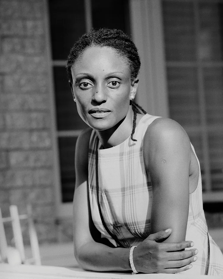 Un portrait en noir et blanc d'une femme afro-américaine avec cheveux en dreadlocks, portant une robe à carreaux, assise avec ses bras croisés sur une surface, dans un décor intérieur.