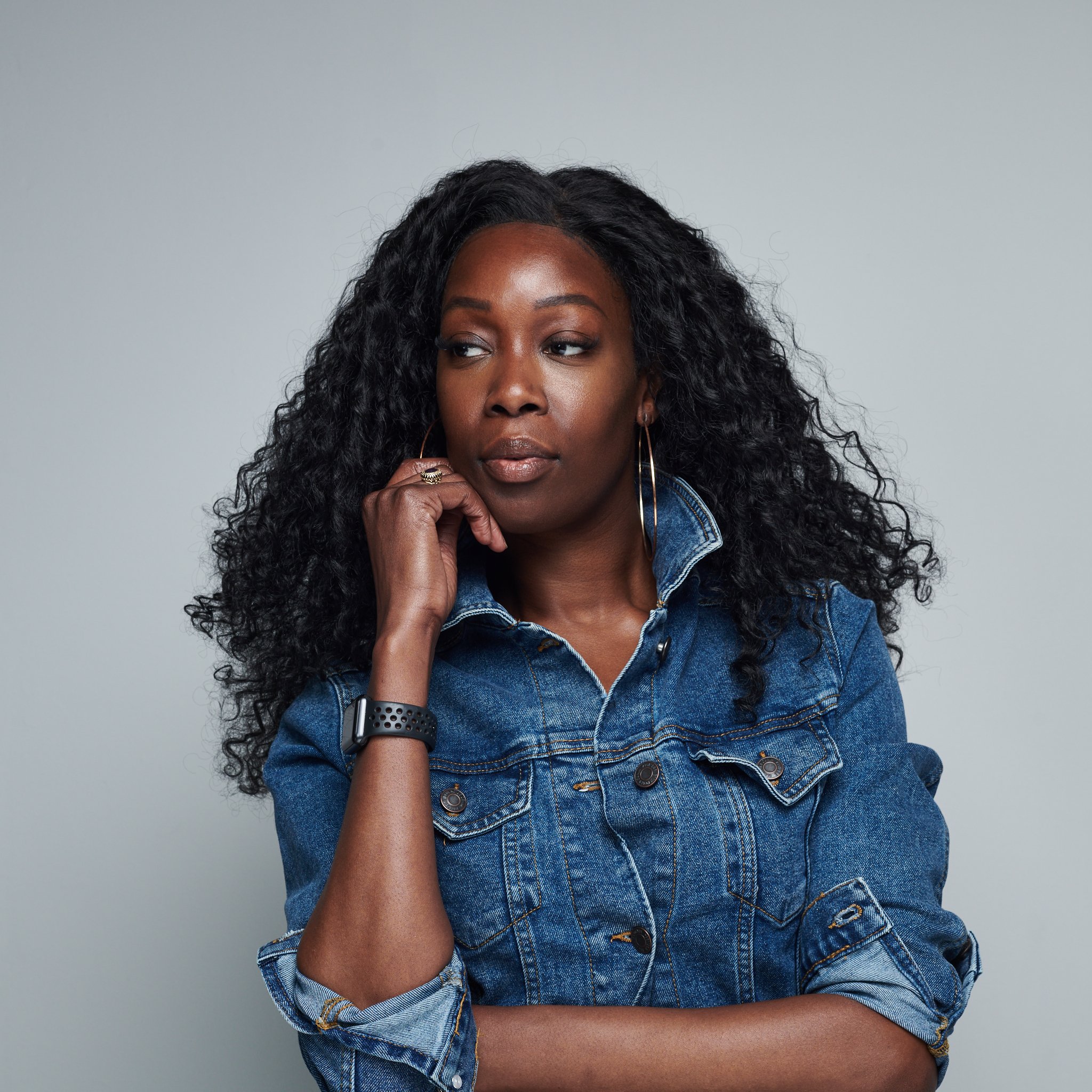 Portrait d'une femme noire avec longs cheveux bouclés noirs portant une veste en jean, portant des boucles d'oreilles en boucle d'oreille et un bracelet, regardant légèrement sur le côté, avec un fond gris clair.