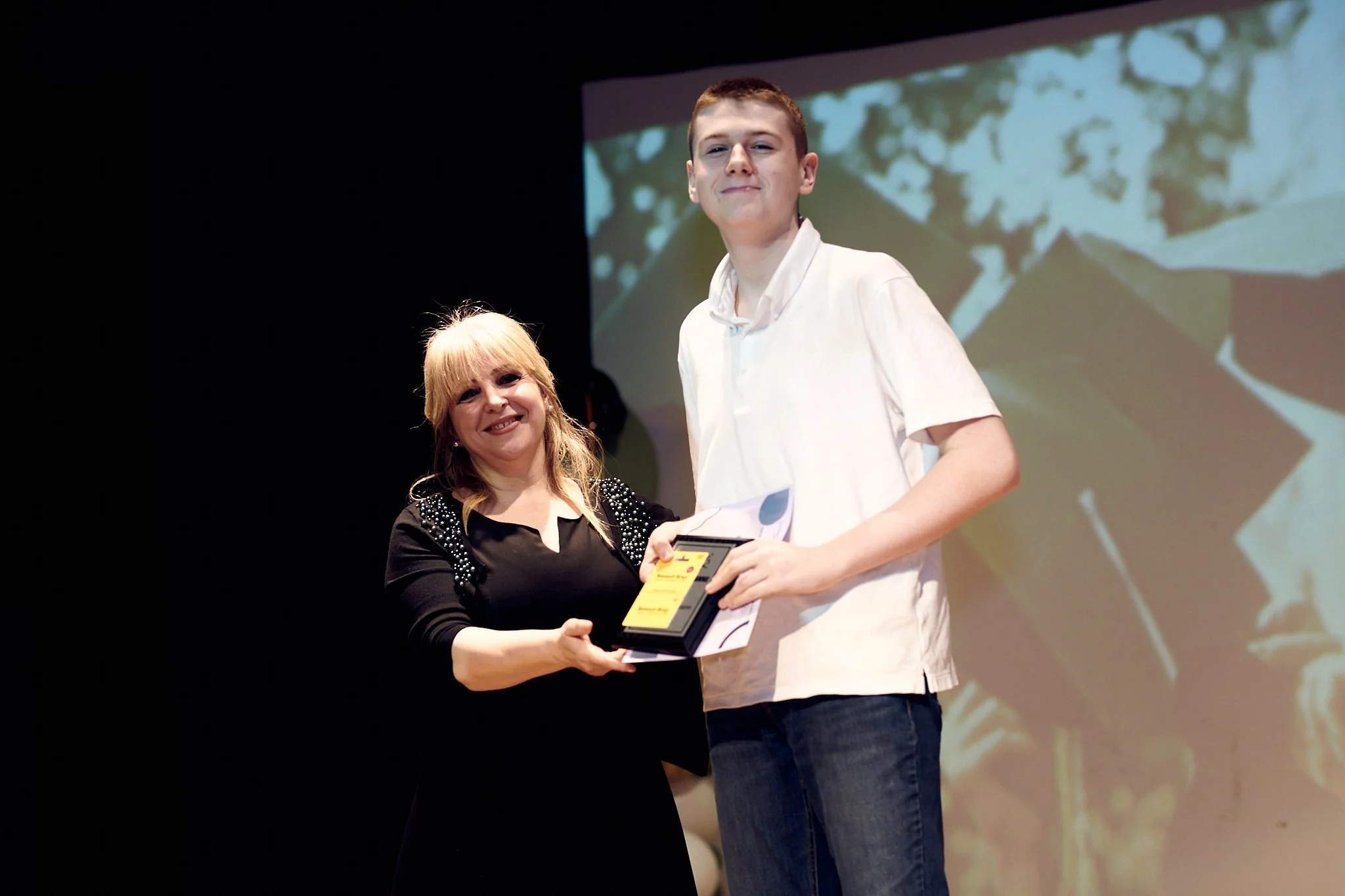 Une femme et un jeune homme souriants lors d'une remise de prix, en train de se serrant la main, avec un écran en arrière-plan.