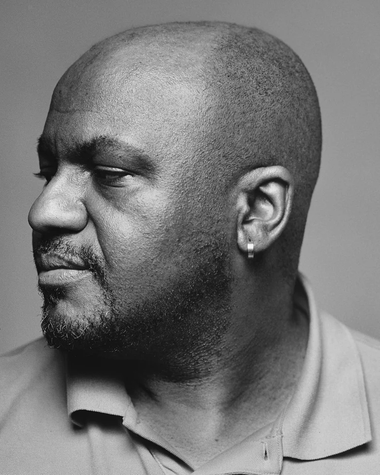 Portrait en noir et blanc d'une personne portant un T-shirt à col rond. La personne a une peau foncée, une barbe et une boucle d'oreille.