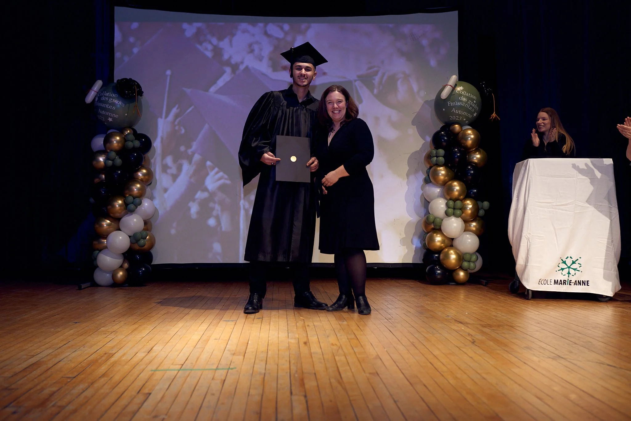 Un étudiant en robe de graduation en train de recevoir son diplôme lors d'une cérémonie à l'école Marie-Anne, organisé avec des décorations de ballons dorés, blancs et noirs.