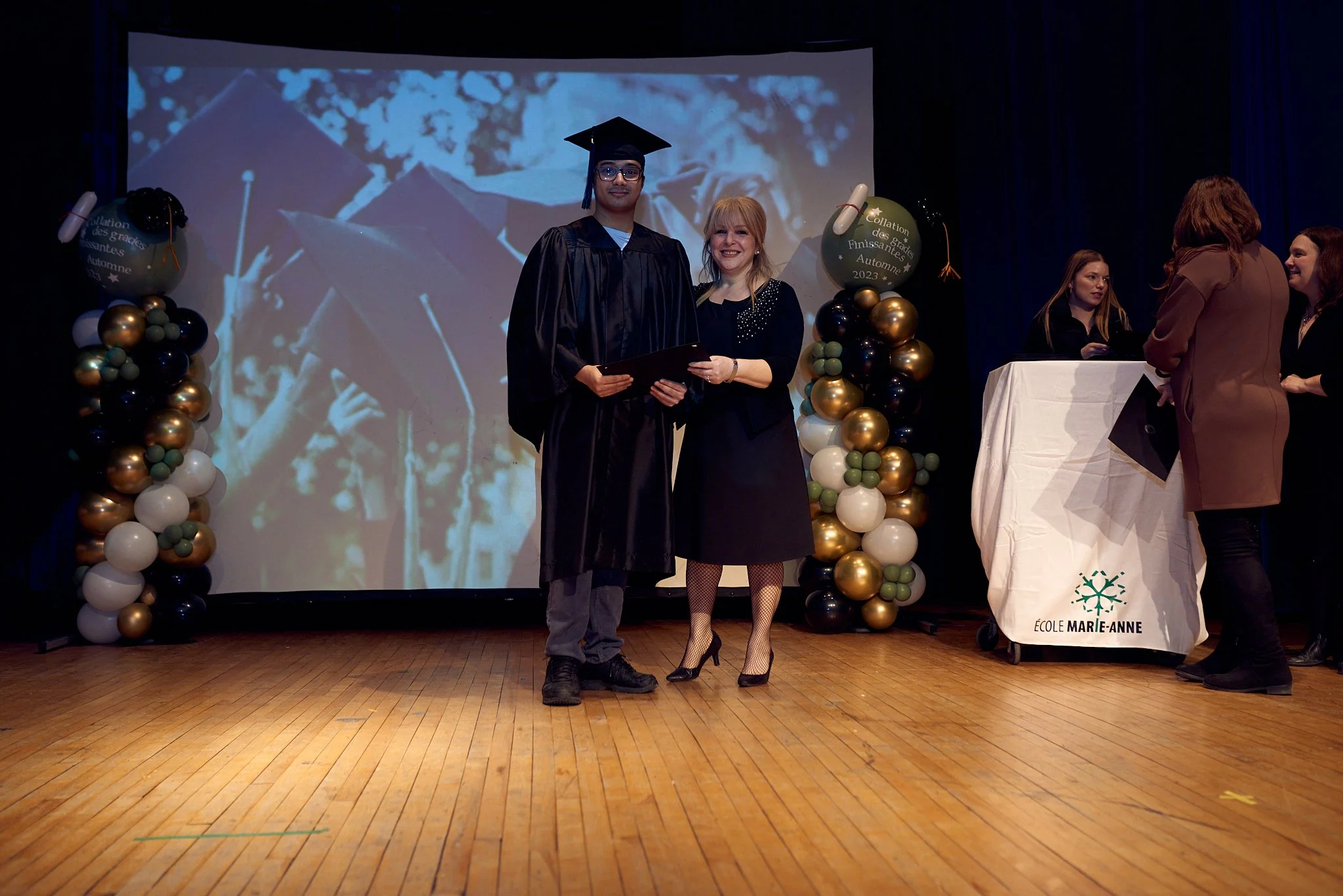 Un diplômé en robe de graduation et un responsable posent ensemble sur scène lors d'une cérémonie de remise de diplômes, décorée avec des ballons dorés, noirs et blancs, devant un grand écran
