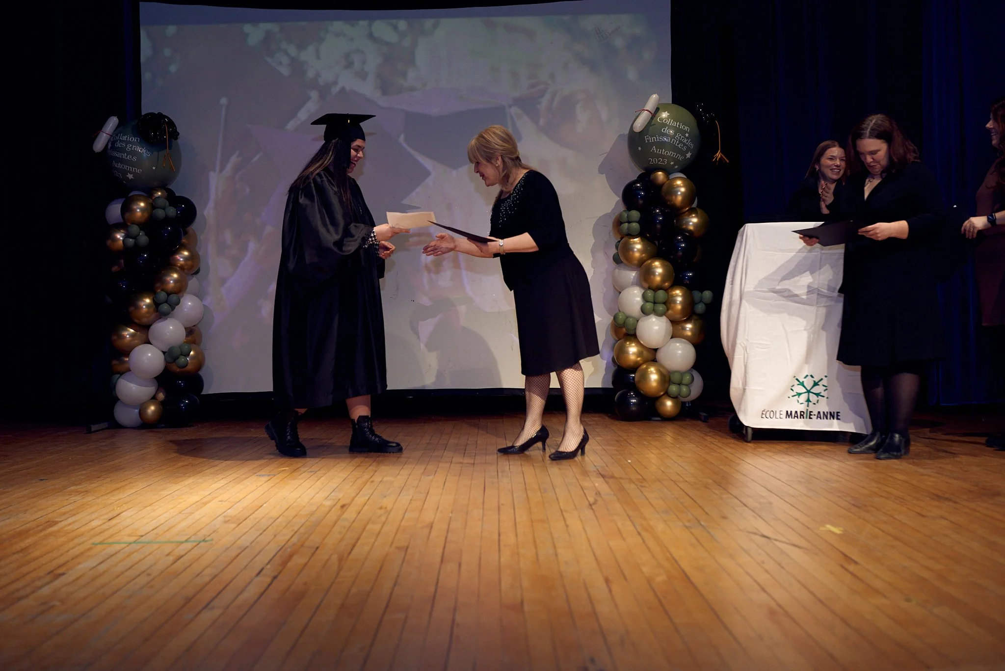 Une cérémonie de remise de diplôme sur scène avec des décorations de ballons, un écran en arrière-plan, une femme en robe de graduation recevant un diplôme d'une autre femme en robe noire, trois autres femmes regardant et tenant des documents.