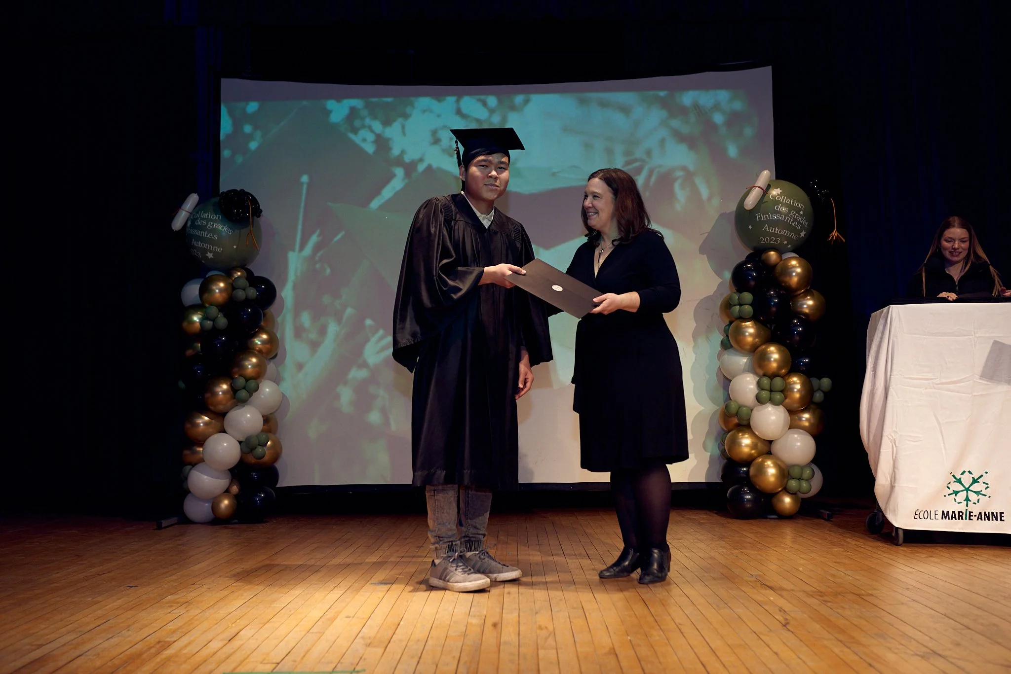 Un étudiant vêtu d'une robe de graduation reçoit un diplôme d'une femme lors d'une cérémonie de remise de diplômes, avec des décorations de ballons dorés, noirs et blancs et une projection en arrière-plan.