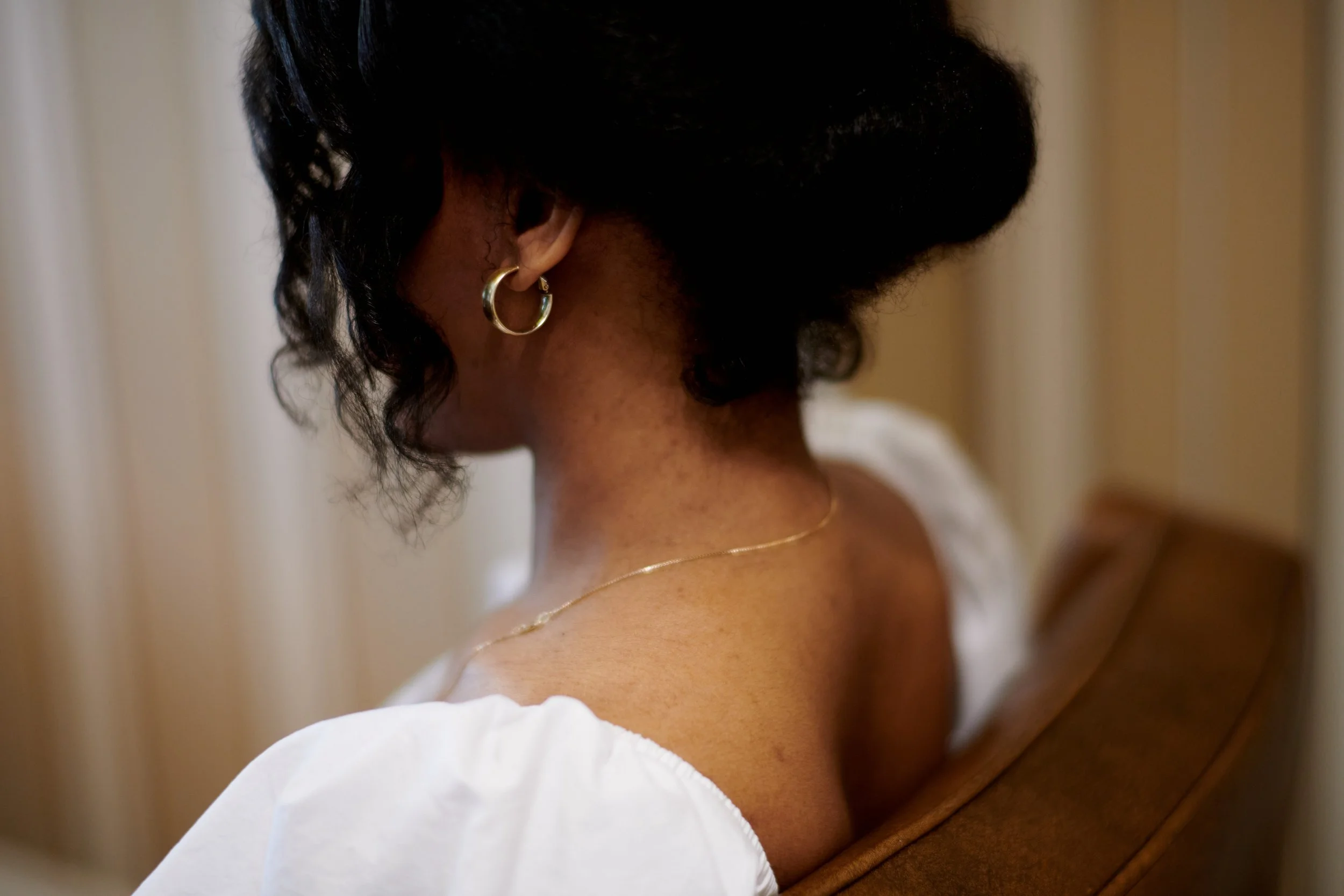 Portrait d'une femme vue de dos, portant des boucles d'oreilles en or et un collier fin, avec une coiffure noire bouclée. Elle est vêtue d'un vêtement blanc et assise sur une chaise en bois, dans un décor intérieur avec des rideaux en arrière-plan.
