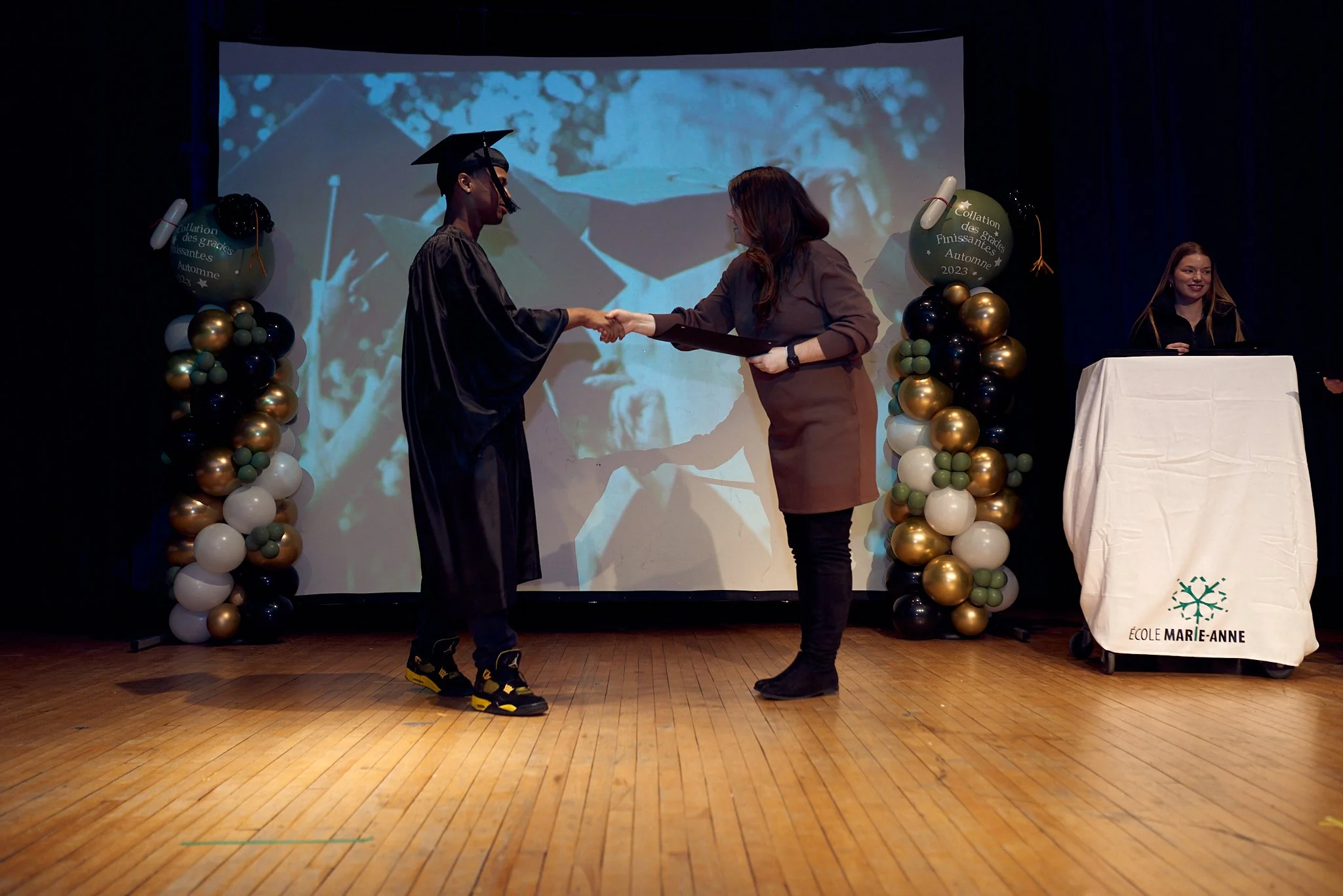 Étudiant diplômé en tenue de graduation laissant la main à un adulte lors d'une cérémonie de remise de diplôme à l'école Marie-Anne, avec des ballons décoratifs et une projection en arrière-plan.