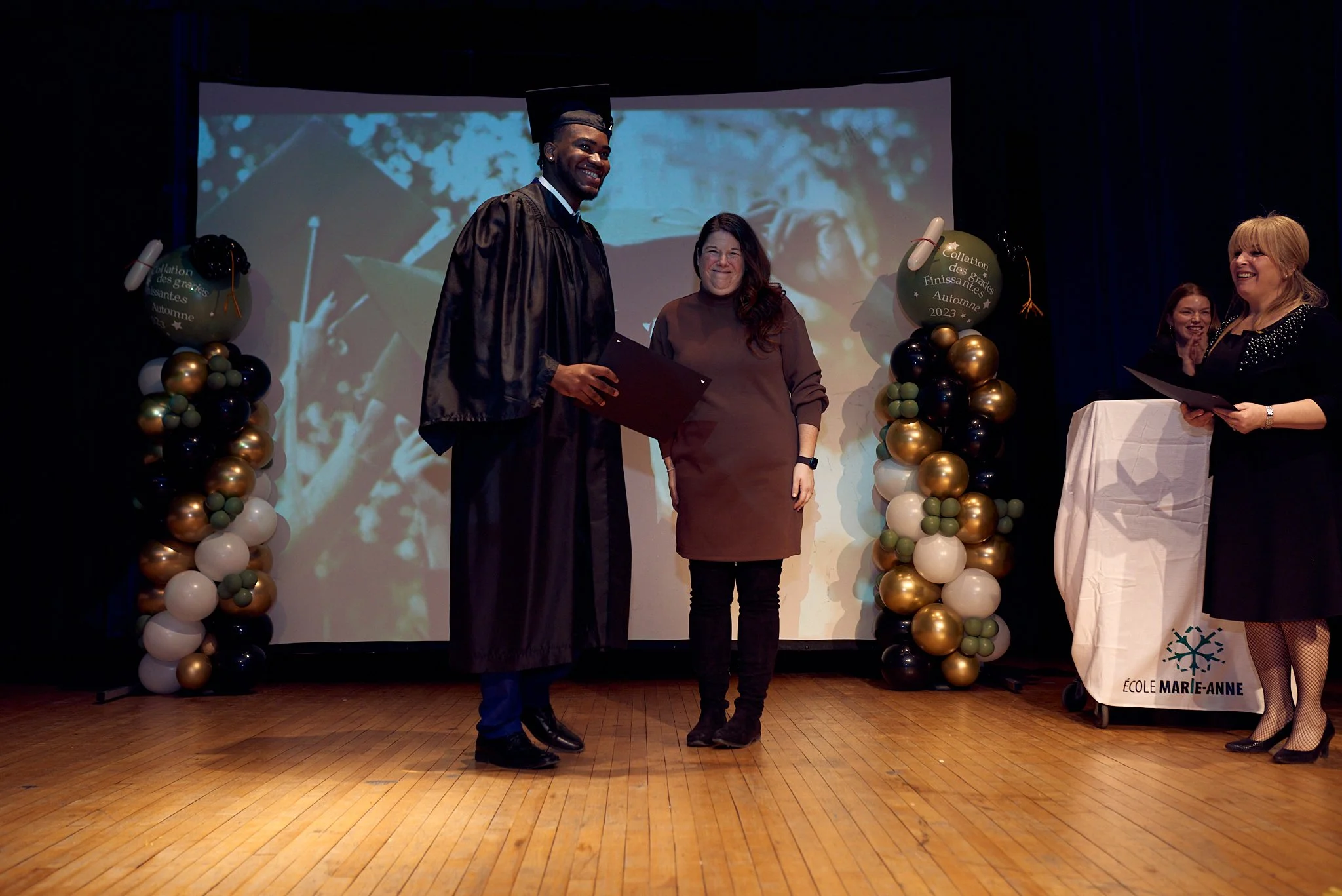 Cérémonie de remise de diplôme lors d'une graduation à l'école Marie-Anne, avec un étudiant en costume de graduation recevant son diplôme sur scène, entouré de décoration de ballons et de personnes applaudissant.