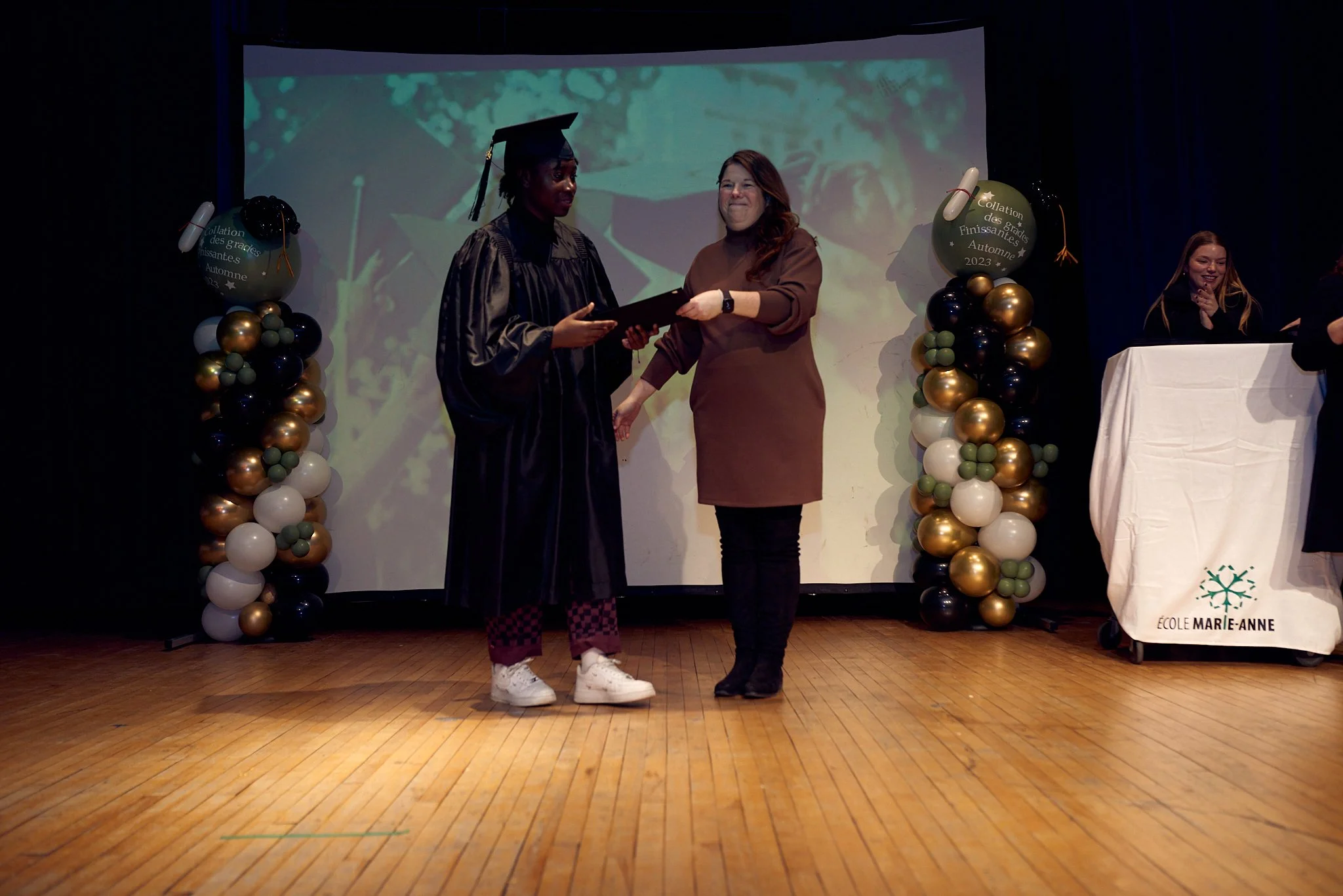 Une cérémonie de remise de diplôme ou de certificat, avec une femme en robe de graduation noire recevant un diplôme d'une autre femme habillée en marron, sur scène décorée avec des ballons noirs, blancs, dorés et verts, lors d'un événement à l'école 