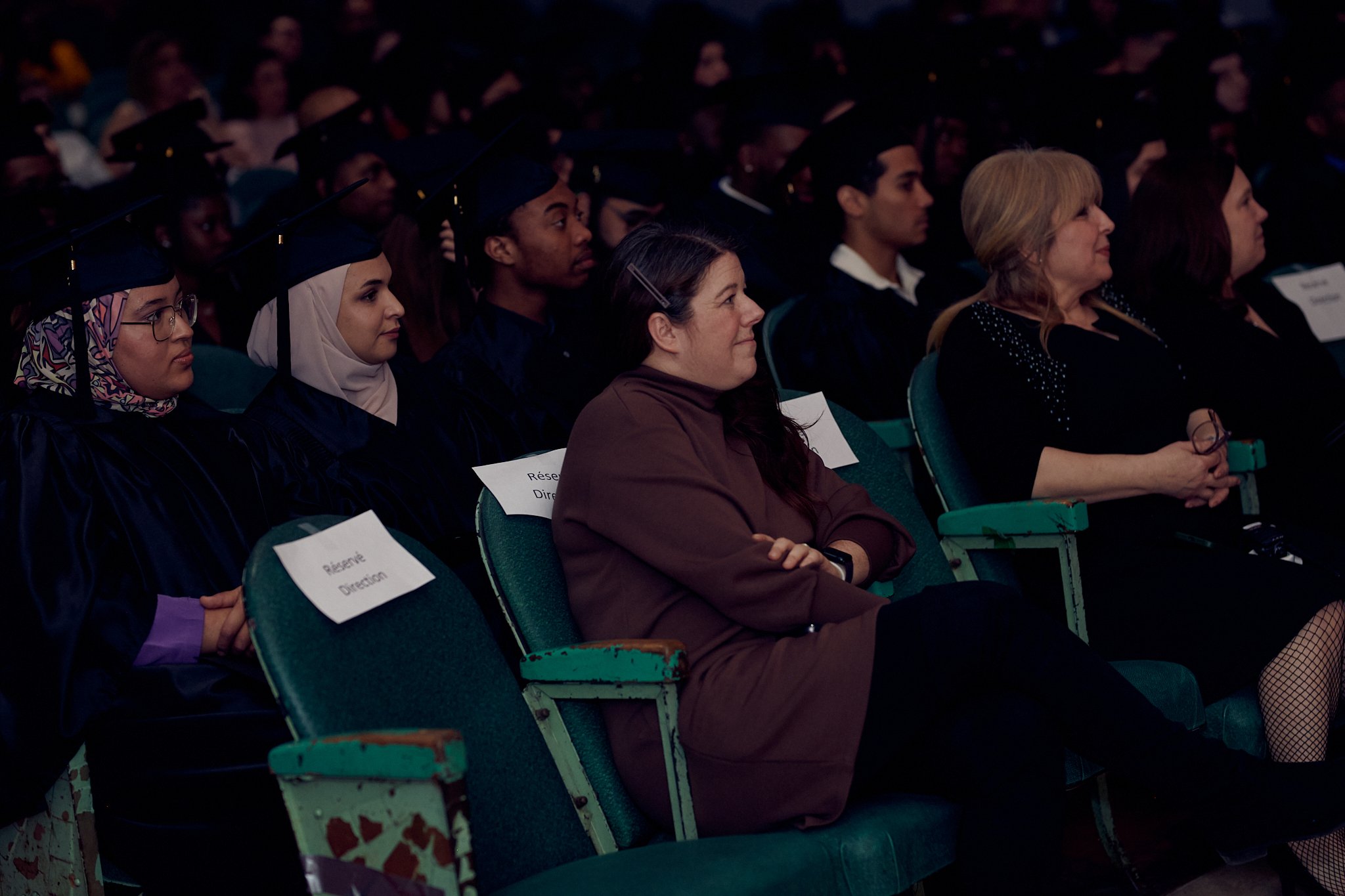 Étudiants en robe de graduation assis dans une salle, certains portant des robes et chapeaux de cérémonie, lors d'une cérémonie de remise de diplômes.