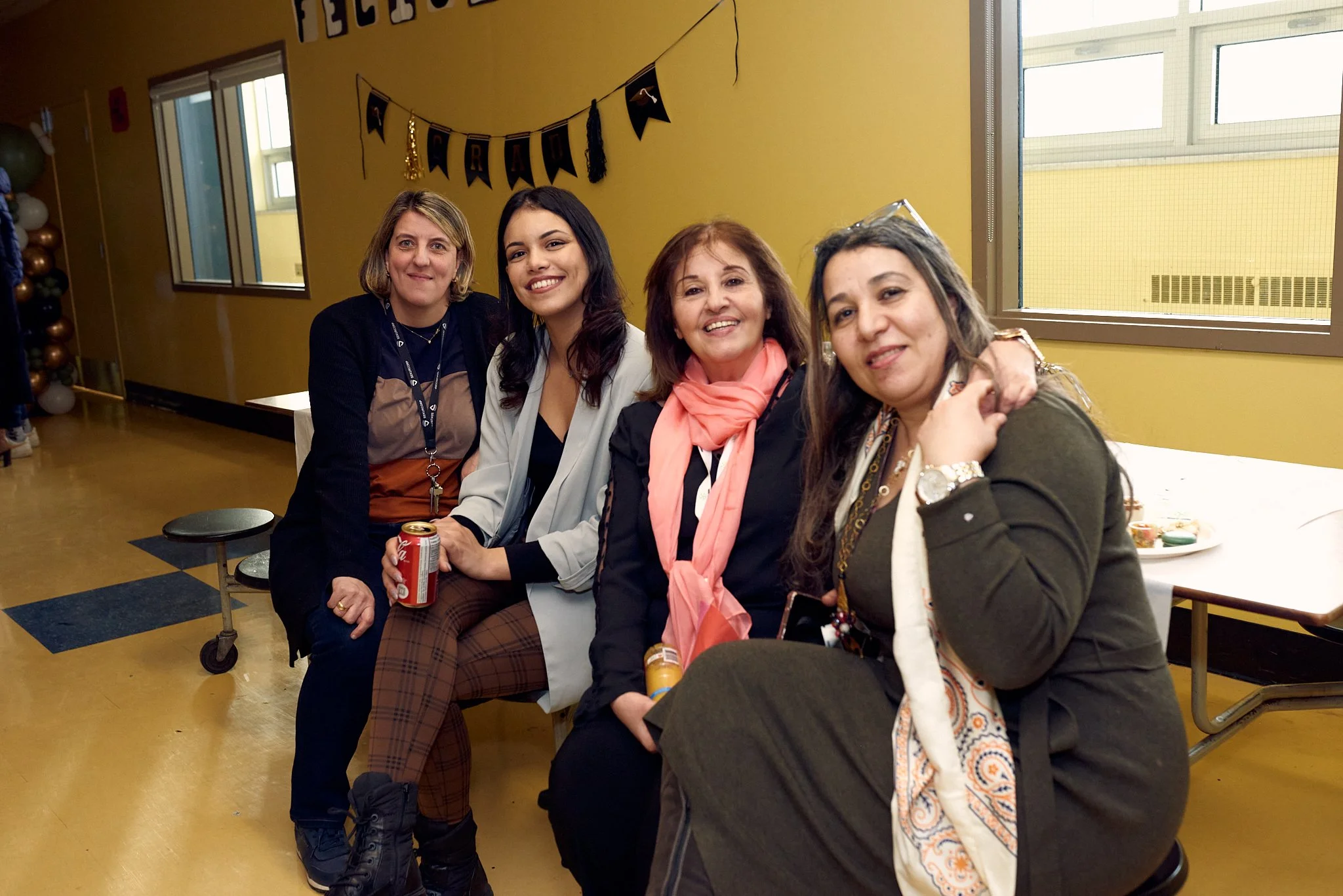 Groupe de quatre femmes assises ensemble, souriantes, dans une salle décorée, probablement lors d'une fête ou d'un rassemblement social, avec des ballons et une bannière.