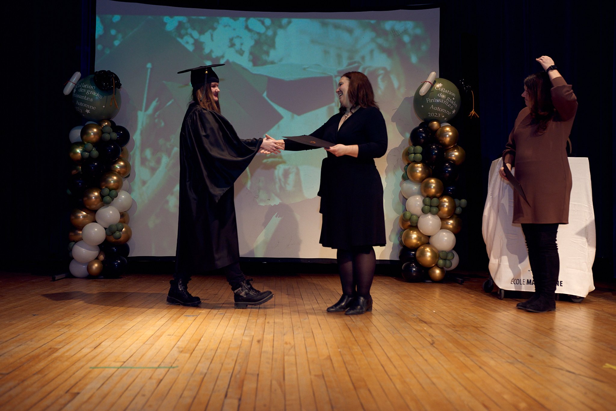 Une cérémonie de remise de diplôme avec trois femmes sur scène, l'une portant une robe de graduation et le chapeau, échangeant un diplôme avec une autre femme. Ballons décoratifs dorés, noirs, et blancs encadrent la scène. Une présentation se déroule