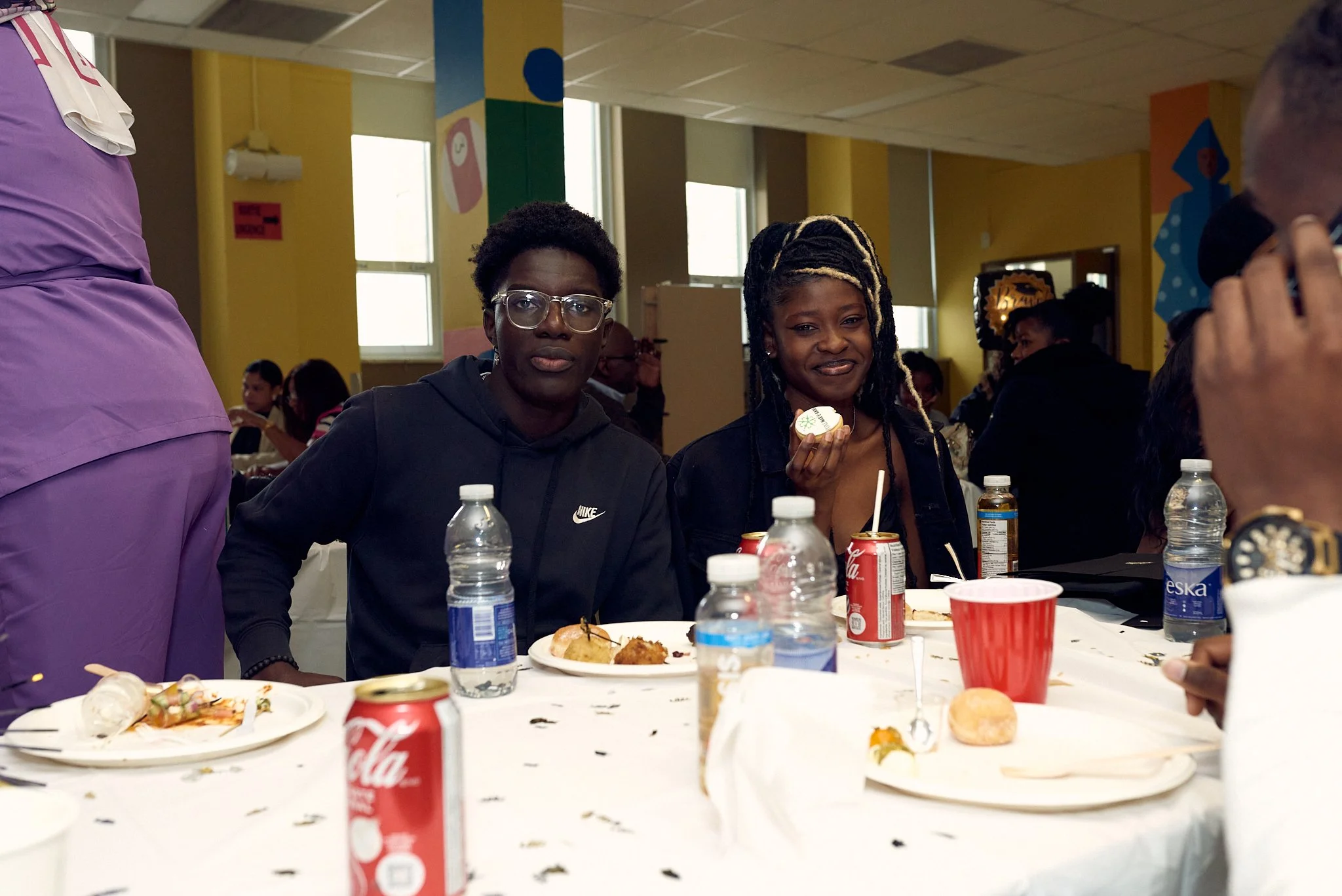 Deux femmes assises à une table lors d'un repas, entourées de bouteilles d'eau, de canettes de Coca-Cola, et de plats, dans une salle décorée colorée.