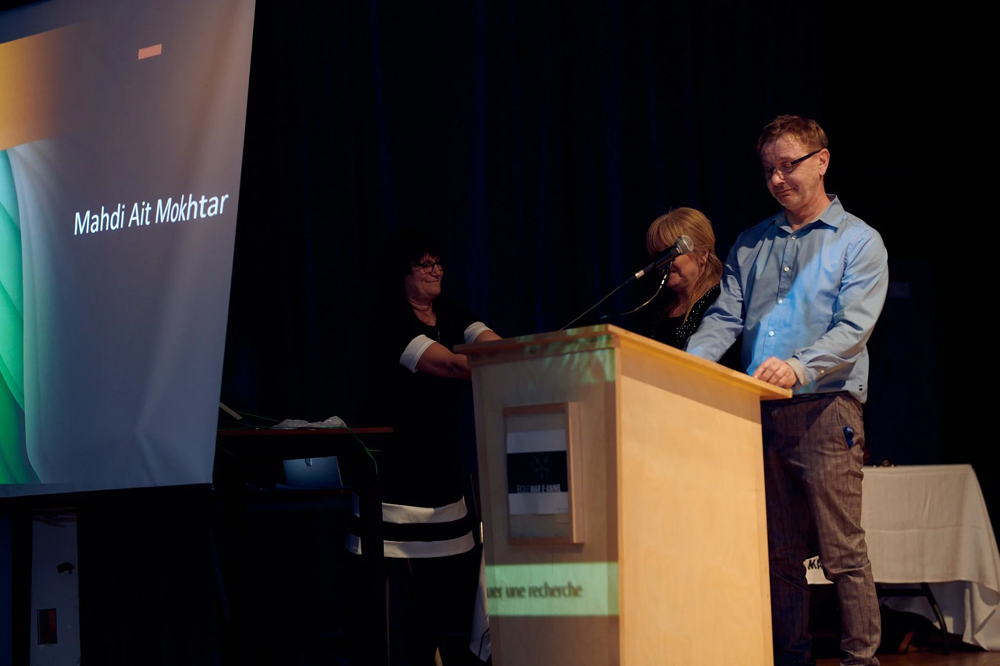 Un homme et une femme se tiennent derrière un pupitre sur scène lors d'une cérémonie ou d'une conférence, une autre femme se tient en arrière, souriante.