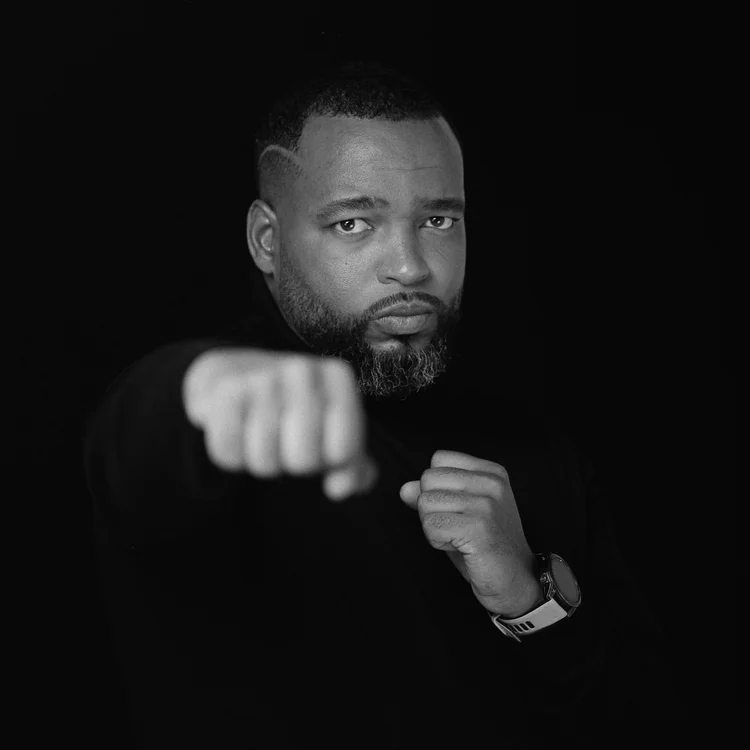 Portrait en noir et blanc d'un homme avec une barbe, portant une montre, faisant un geste de poing fermé avec la main. Photo prise en gros plan contre un fond noir.