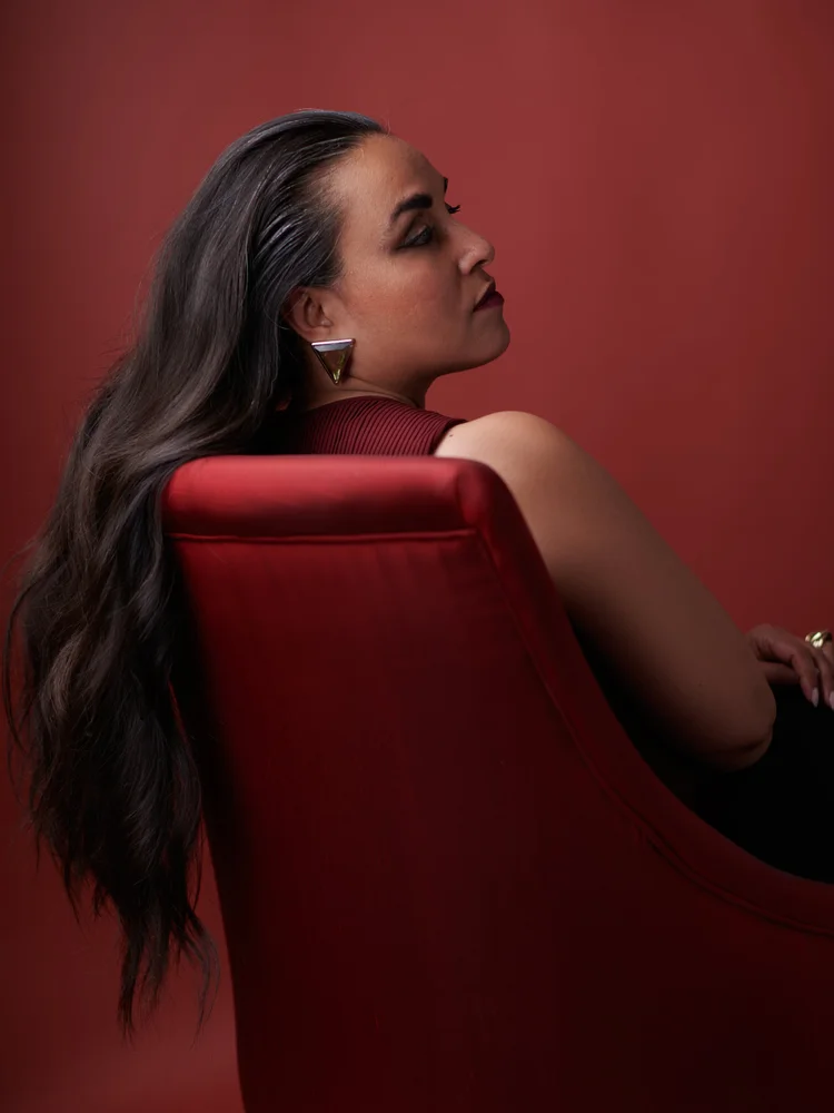 Femme aux longs cheveux ondulés, assise de dos sur un fauteuil rouge, regardant par-dessus son épaule, boucle d'oreille argentée, fond rouge brique.