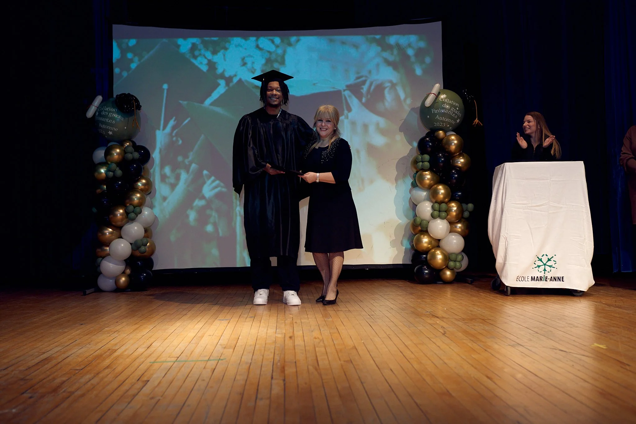Cérémonie de remise de diplôme sur scène, avec un jeune diplômé en costume de graduation recevant un prix, entouré de décorations de ballons et d'un écran en arrière-plan.