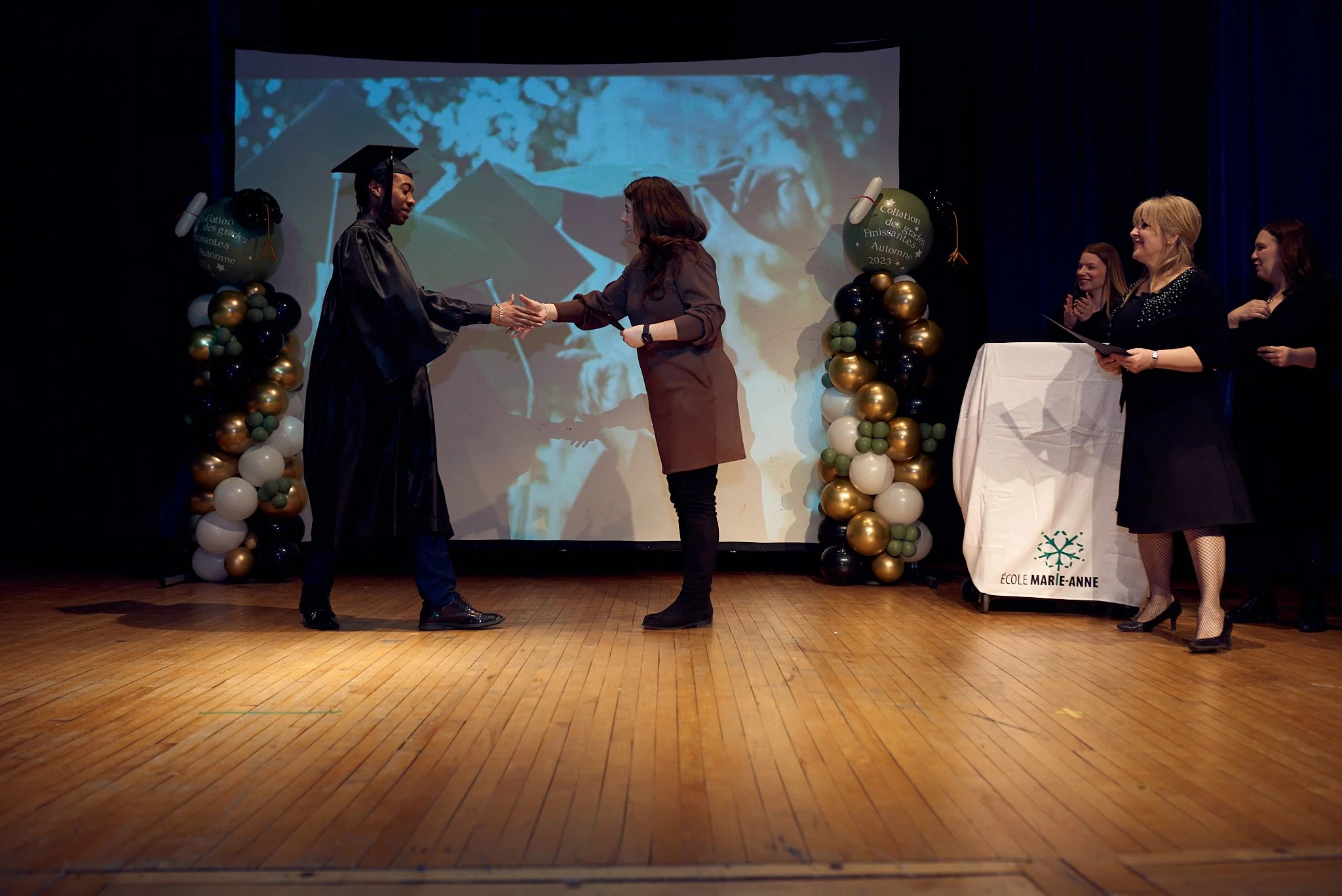 Une cérémonie de remise de diplôme avec une personne en costume de graduation recevant son diplôme d'une femme sur scène, entourée de trois autres femmes applaudissant, avec des décorations de ballons et un fond vidéo.