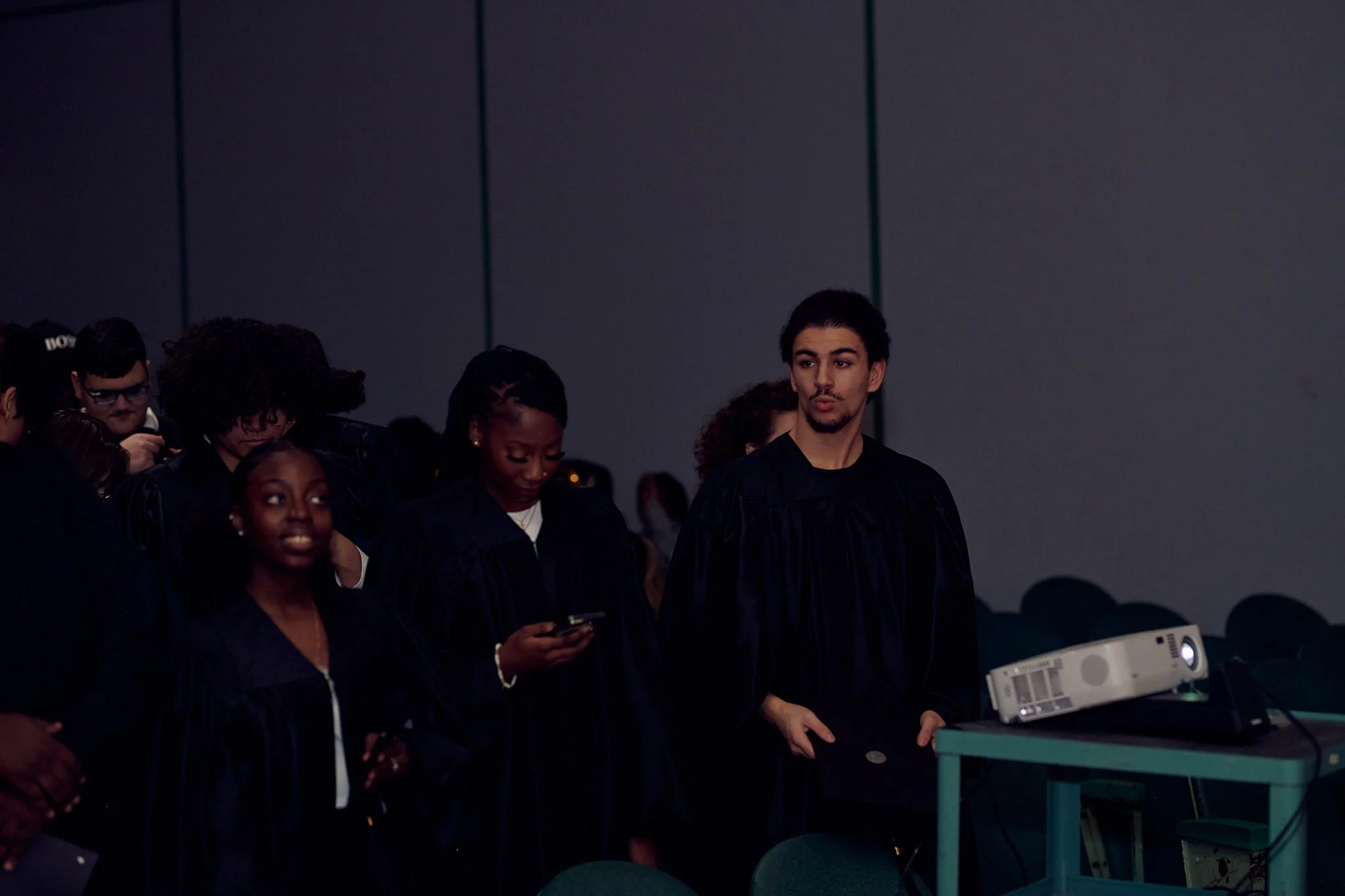 Groupe de jeunes gens vêtus de robes de graduation dans une salle sombre, certains regardant leurs téléphones ou une présentation projection.