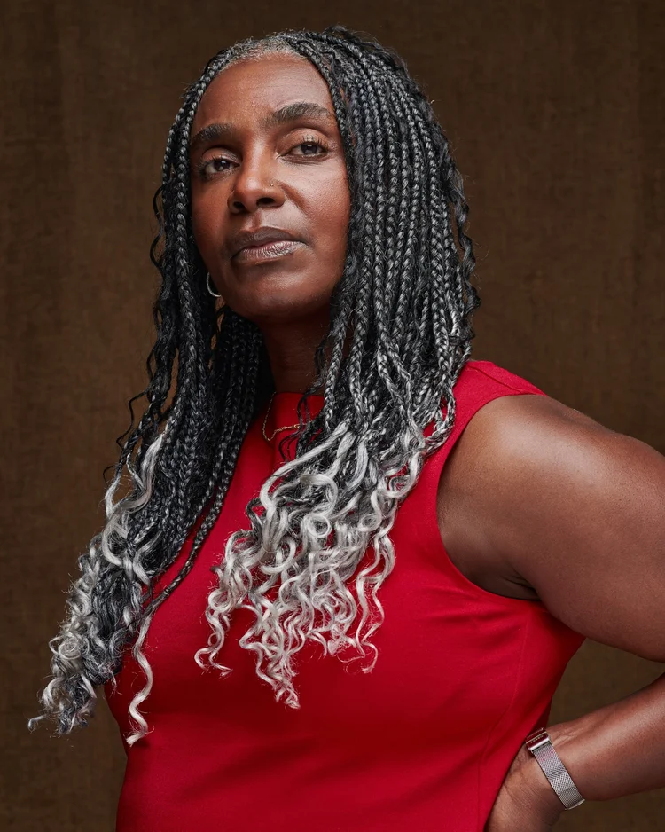 Une femme noire aux cheveux gris et bouclés, portant un haut rouge, pose devant un fond marron.