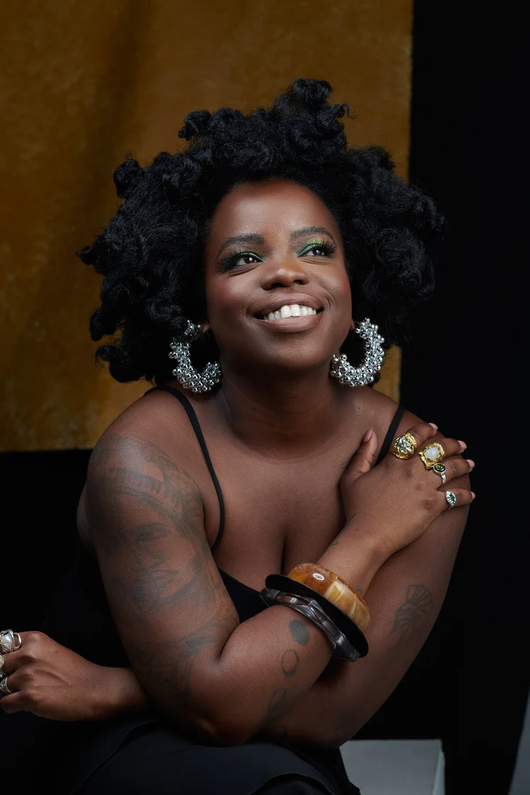 Un portrait d'une femme noire avec des cheveux crépus en bouquet, portant de grosses boucles d'oreilles en perles, plusieurs bagues et bracelets, portant un haut noir à fines bretelles. Elle sourit et regarde vers la droite, avec un fond noir et or.