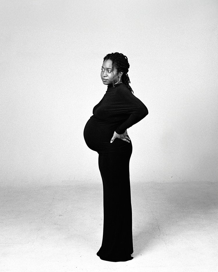 Une femme enceinte portant un manteau noir se tient la main sur la hanche, regardant vers la gauche, devant un mur blanc.