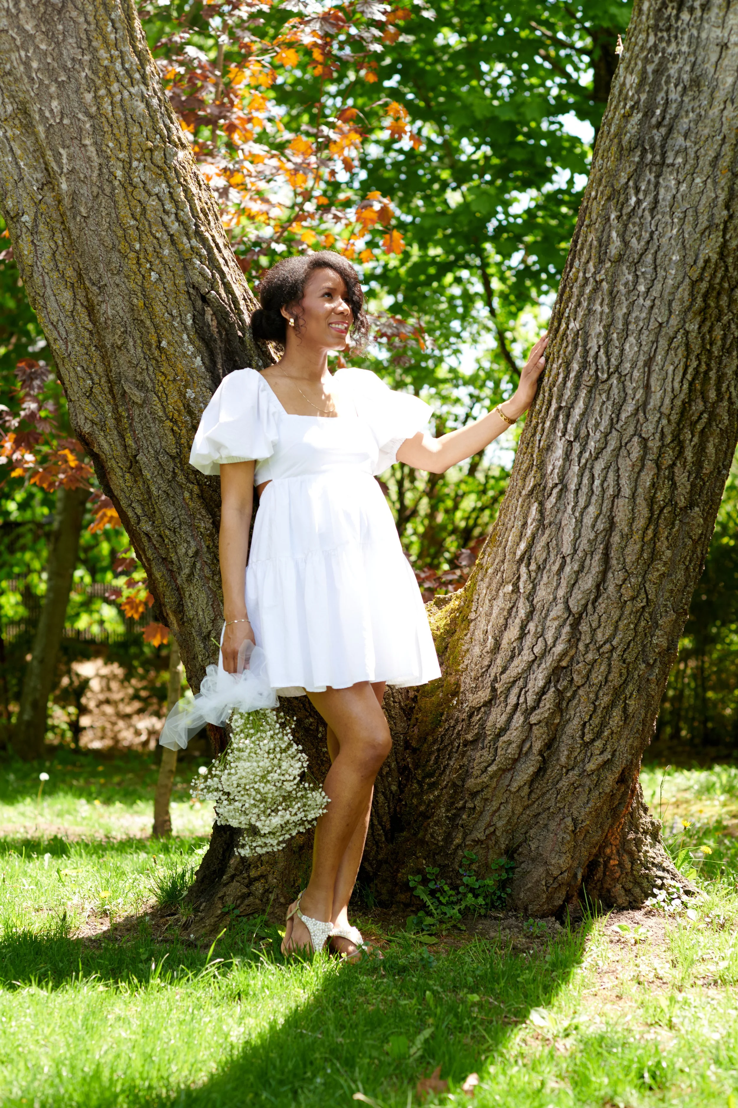 Une femme en robe blanche souriante, posant dans un parc sous un arbre, tenant un bouquet de fleurs blanches.