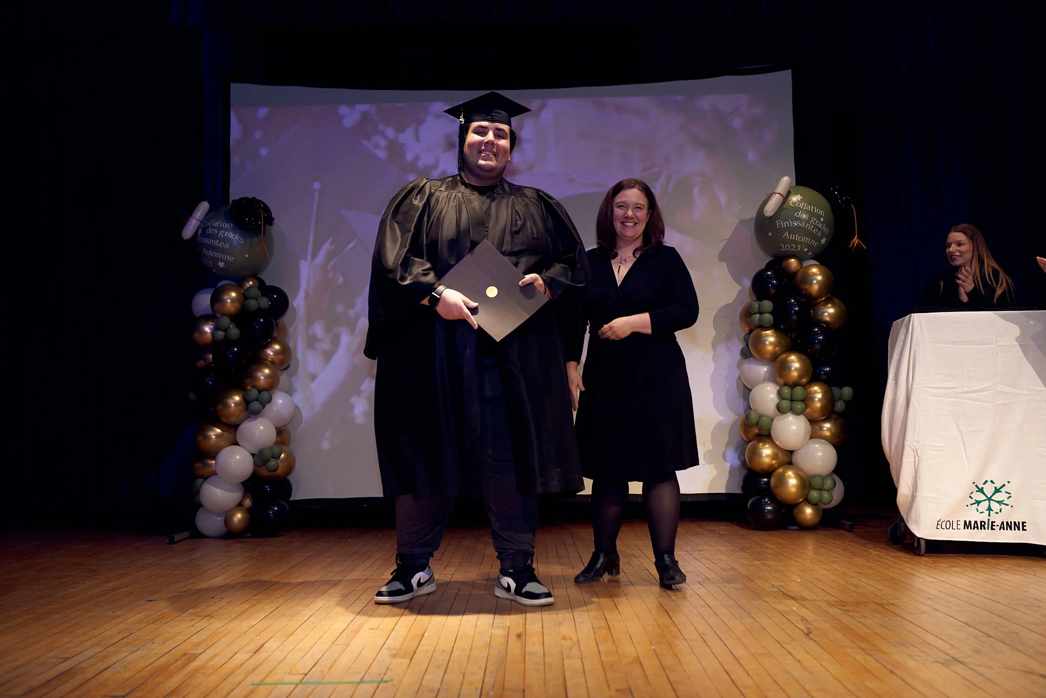 Un étudiant en costume de graduation tenant un diplôme, posant avec une femme lors d'une cérémonie de remise de diplômes à l'école Marie-Anne.