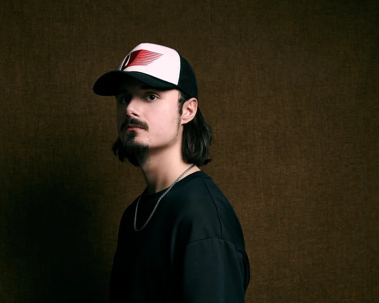 Un jeune homme porte une casquette blanche et noire avec un logo rouge, un collier argenté, et porte un t-shirt noir sur un fond brun
