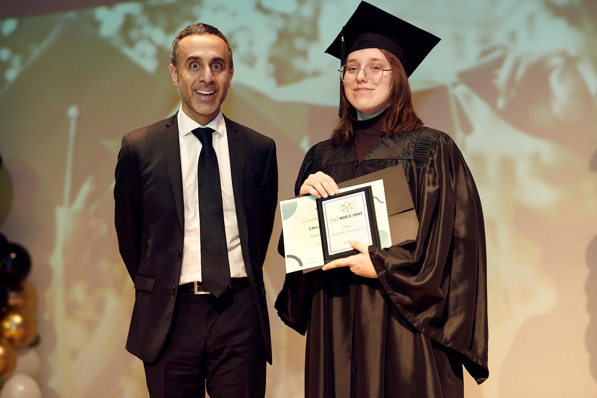 Une jeune diplômée en robe de graduation reçoit un prix lors d'une cérémonie, tenue d'un chapeau de diplômée, en compagnie d'un homme en costume à gauche, lors d'une cérémonie de remise de diplômes.