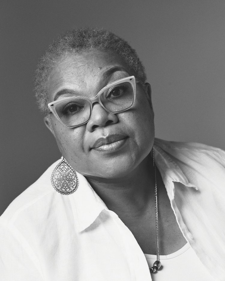 Portrait d'une femme âgée portant des lunettes épaisses, des boucles d'oreilles en dentelle, une chemise blanche avec un col, un collier avec des perles et une chaîne en métal, dans une photo monochrome en noir et blanc.