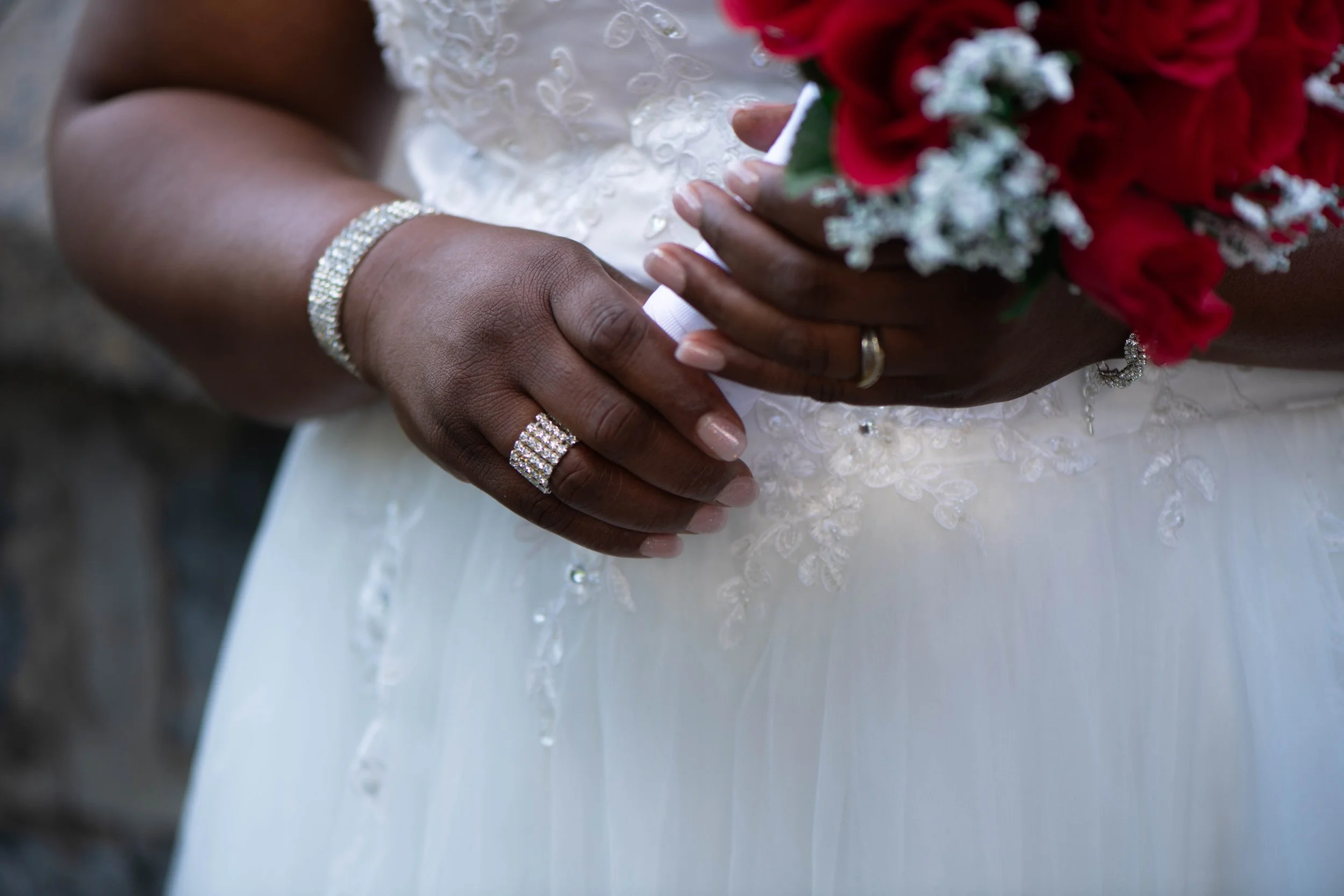 Close-up des mains d'une mariée portant des alliances et des bijoux, tenant un bouquet de fleurs rouges et blanches, vêtue d'une robe de mariage blanche avec des détails en dentelle.