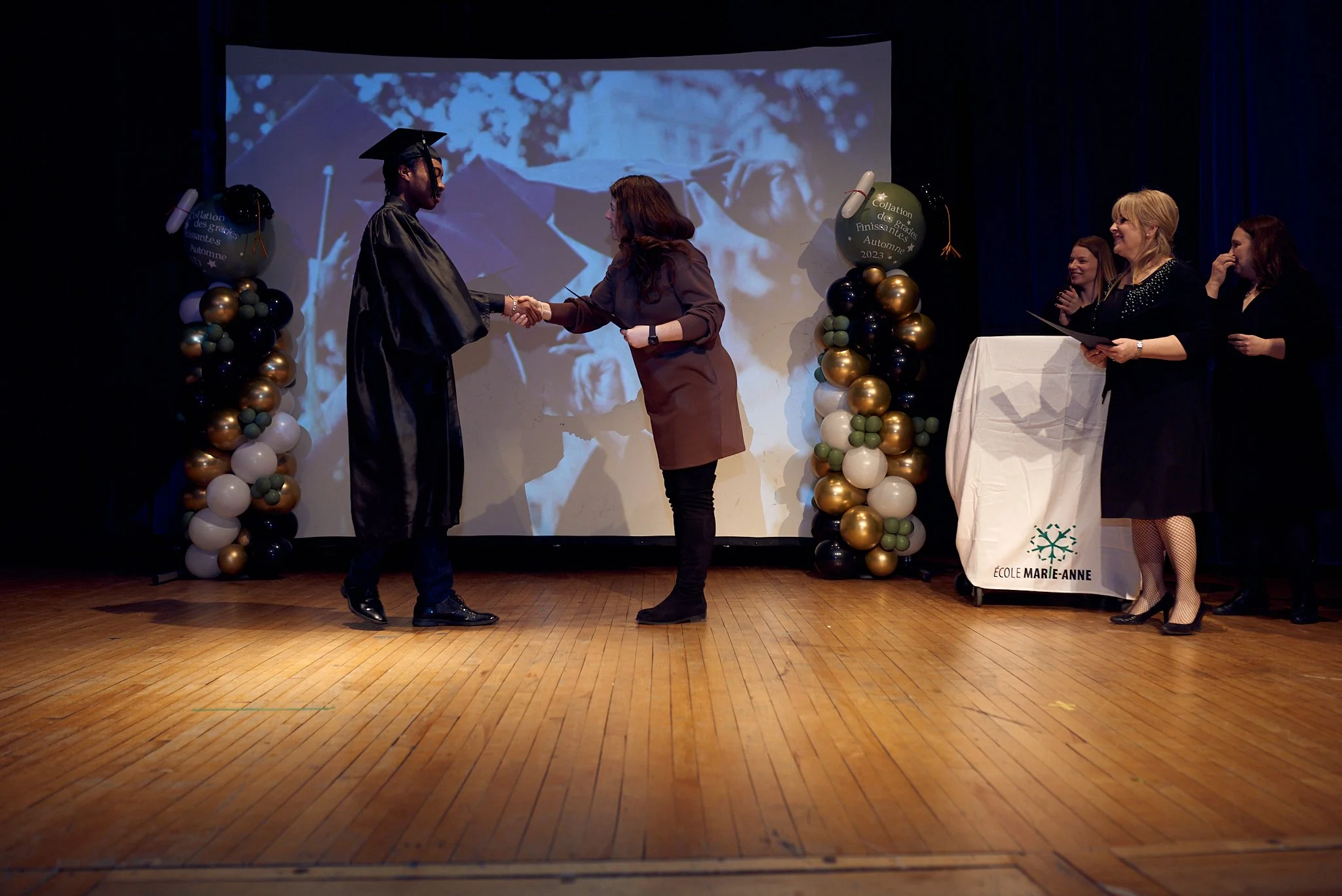 Cérémonie de remise de diplôme lors d'une graduation à l'école Marie-Anne, avec un étudiant en robe de graduation recevant son diplôme d'une femme, entourée d'autres personnes sur scène.