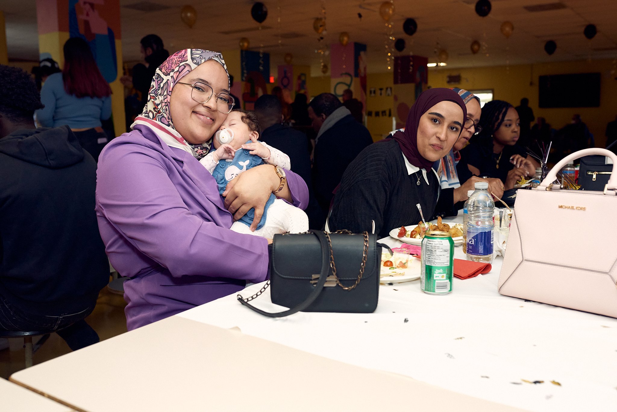 Groupes de femmes et d'enfants assis à une table lors d'un repas ou d'une fête dans une salle colorée décorée de ballons et de guirlandes.