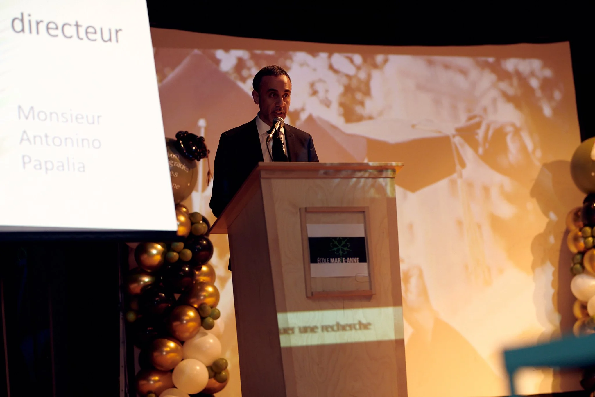Un homme en costume noir parle derrière un podium en bois lors d'une réunion ou d'une cérémonie, avec un écran projecteur à gauche affichant du texte en français et un décor de ballons dorés, blancs et verts décorant le côté gauche de la scène.