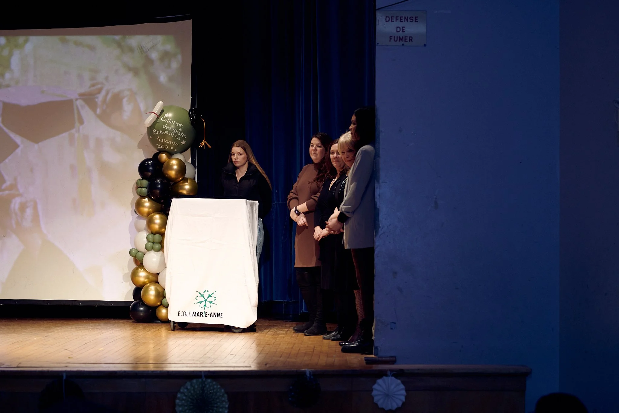 Quatre femmes debout sur une scène d'école lors d'une cérémonie, avec un tableau blanc, un ballon et une bannière "École MARIE-ANNE" visible, dans une salle avec un rideau bleu