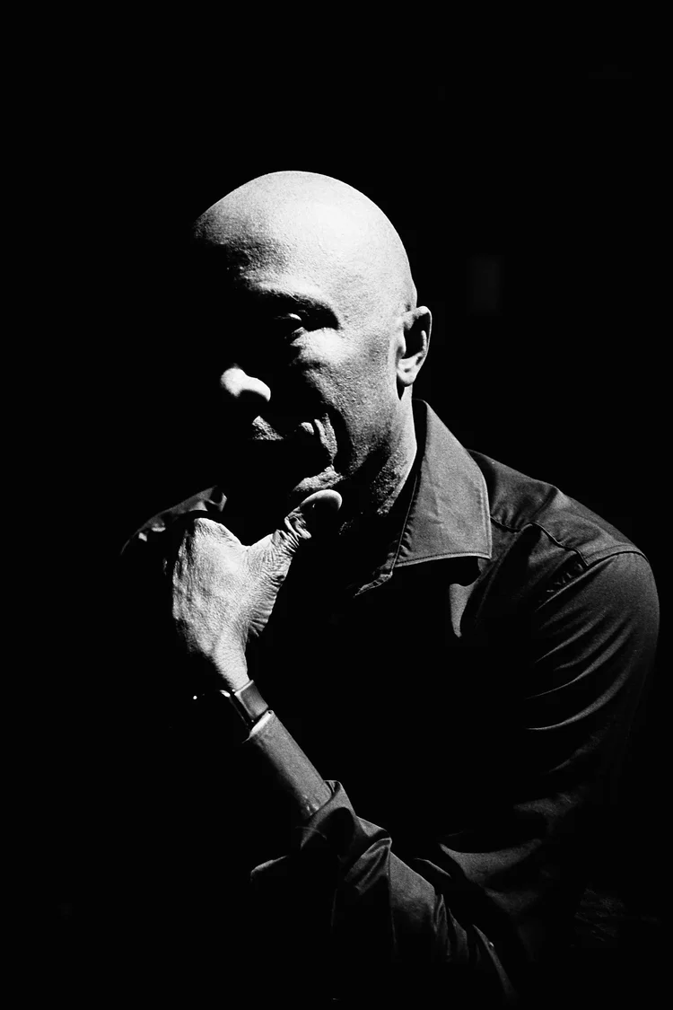 Portrait dramatique en noir et blanc d'un homme chauve, la main sur le menton, éclairage contrasté sur fond sombre.