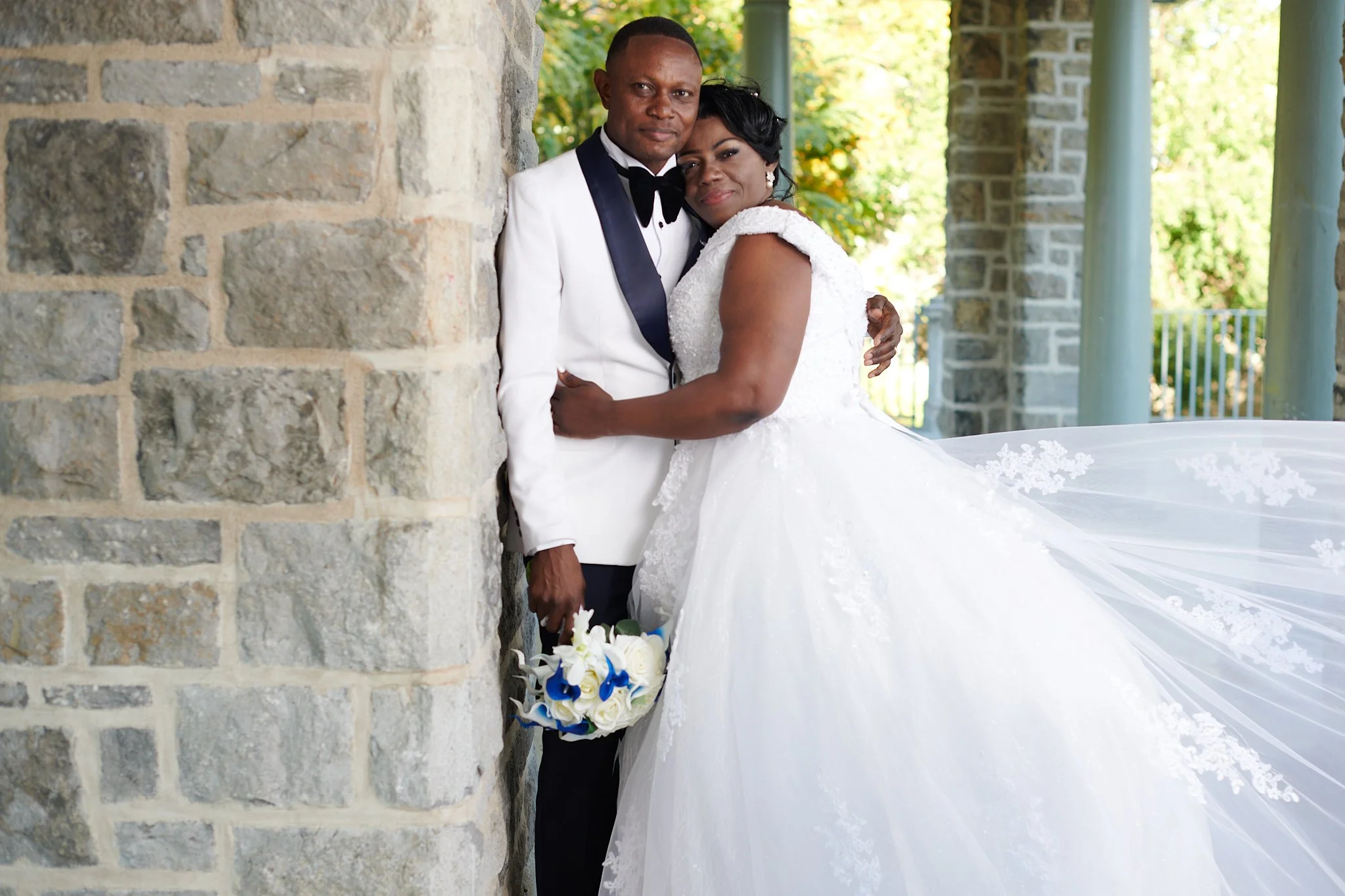 Un couple en vêtements de mariage posant sous une structure en pierre, la femme en robe blanche et le homme en smoking blanc, avec un bouquet de fleurs.