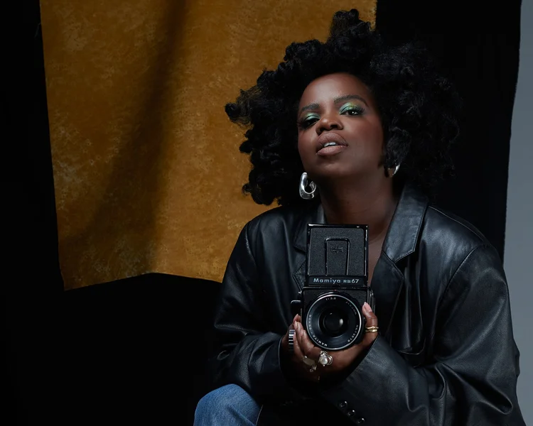 Selfie d'une femme noire avec des cheveux bouclés noirs, portant un maquillage vert et des boucles d'oreilles en forme de cœur, tenant un appareil photo Mamiya RB67, habillée en cuir noir, en face d'un fond coloré